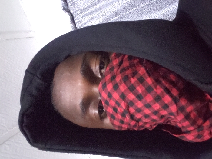 A person wearing a black hoodie and a red and black checkered bandana covering the lower half of their face. Their eyes are visible above the bandana. The background includes a textured white wall and a gray, possibly metallic, surface. The person's skin tone is dark. The image appears to have been taken from a slightly high angle. The photograph is slightly rotated.