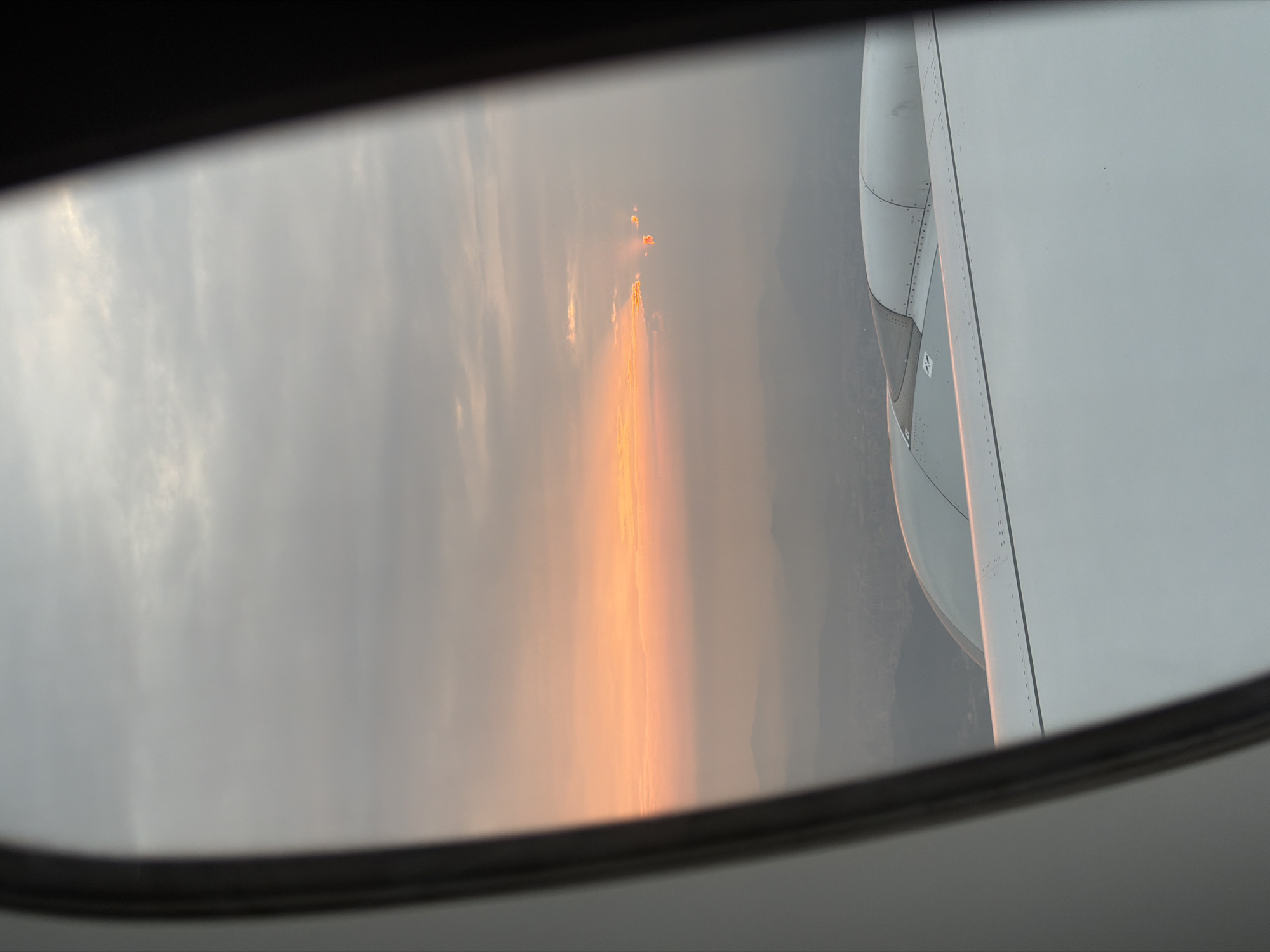 The image, taken from an airplane window, captures a dramatic sky scene, likely at sunrise or sunset, above the Baiyun District, China. The dark frame of the aircraft window outlines the view. To the right, a significant part of the airplane's wing and engine cowling is visible, detailed with rivets and panel lines in shades of grey and white, subtly reflecting the warm light.
The sky is largely overcast or hazy, with muted grey and white tones in the clouds. In the distance, a faint, darker landmass, consistent with mountains or hills, can be discerned beneath the horizon.
The central focus of the image is a prominent, intense vertical streak of orange and red light, suggesting a powerful light source or reflection. Above this main column, a cluster of a few distinct, bright orange-red lights is visible in the upper mid-distance. The fiery coloration and the distinct vertical emanation strongly suggest a large-scale fire on the ground, possibly a wildfire, with its light and smoke diffused by the atmospheric haze or clouds. There is no clearly legible text visible within the frame.