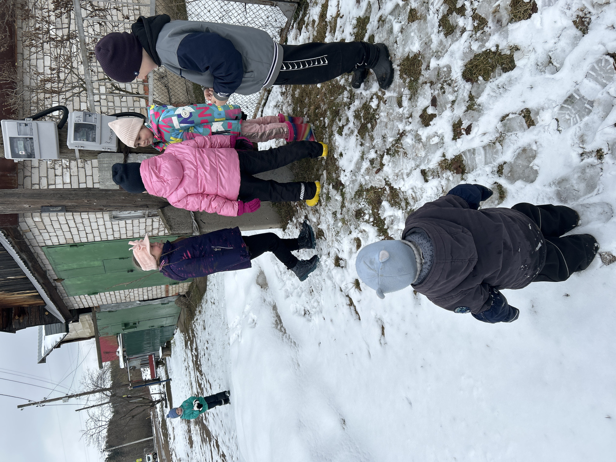 The image depicts a group of children in a snowy outdoor environment. They are located near a row of garages with electrical meters mounted on a brick wall. The scene includes several children in winter clothing, walking, and standing near the garages and snow-covered ground. The sky is overcast, and the environment appears to be in a residential area. This is the city of Chaikovsky, Russia.