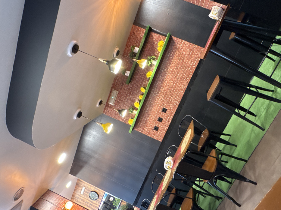 The image displays the unoccupied interior of a commercial establishment, likely a cafe or restaurant, located in Bragança, Portugal. The perspective is an angled, slightly elevated view, looking towards the back and right.
The background features a prominent wall covered in a red-brown faux brick pattern, with a dark, possibly black, wainscoting along its base. Two horizontal, narrow shelves, colored green, are mounted on this brick wall. The lower shelf holds multiple yellow, flower-like decorative items and small potted plants, while the upper shelf similarly displays potted plants and some yellow decorations. Below these shelves, four black electrical outlets or data ports are visible on the brick.
The ceiling is white with a dark grey/black curved architectural feature running horizontally across the upper portion of the frame. Multiple pendant lights, some with yellow shades and others with dark grey or black shades, hang from the ceiling. Recessed lighting is also visible. A section of the left wall, towards the rear, features dark wood-like paneling and includes a circular clock with Roman numerals. The wall closer to the foreground on the left is solid dark grey or black with a textured surface.
The flooring throughout the main seating area is a bright, artificial green, resembling turf or carpet, contrasting with a darker floor section visible in the extreme foreground-left.
Seating arrangements consist of two types. On the left, a long, narrow wooden table with a reddish-orange runner and placemats is paired with multiple black metal chairs featuring wooden seats. A white, patterned ceramic object rests on this table. On the right, two tall, narrow wooden tables are positioned, each accompanied by black metal bar stools with wooden tops. One of these tables also holds a white, patterned ceramic object.
No individuals are present in the scene.