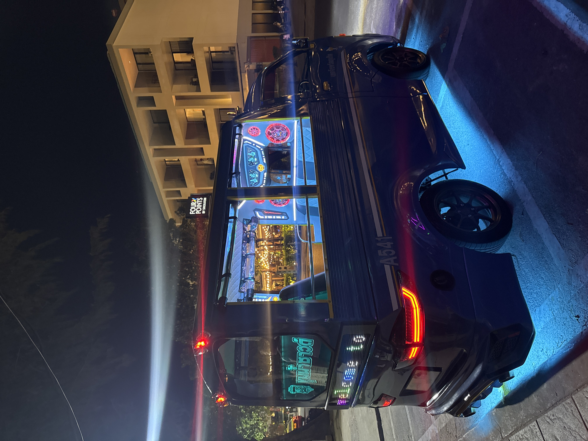 At night in Patong, Thailand, a blue tuk-tuk with custom lighting and sound system is parked on a street. The tuk-tuk has a large sticker on the rear window and lights around the exterior and interior. A building with lit windows is visible in the background, identified by a sign as Four Points by Sheraton. The street has a blue glow.