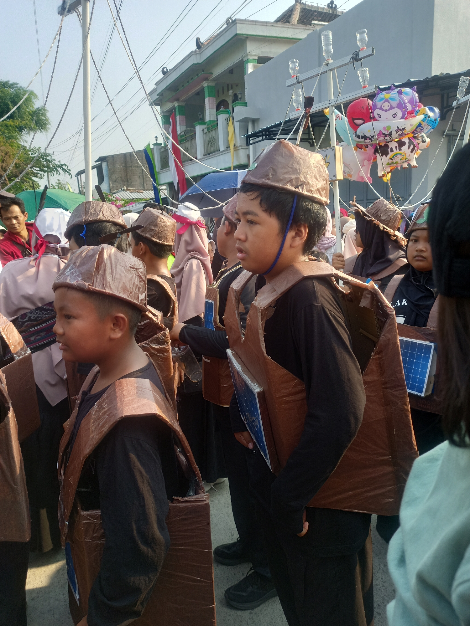 The image depicts a street scene in Gemolong, Indonesia, where a group of children or young people are participating in an event, likely a parade or festival. They are wearing homemade costumes resembling a traditional or religious outfit, with brown cardboard headpieces and chest coverings. Some of the chest coverings have solar panel props attached. The background shows a building with green trim and flags, suggesting it might be a public building or school. In the background, a collection of balloons, including some with cartoon characters, are visible, attached to overhead wires. The atmosphere appears to be bright and sunny, with a clear sky.