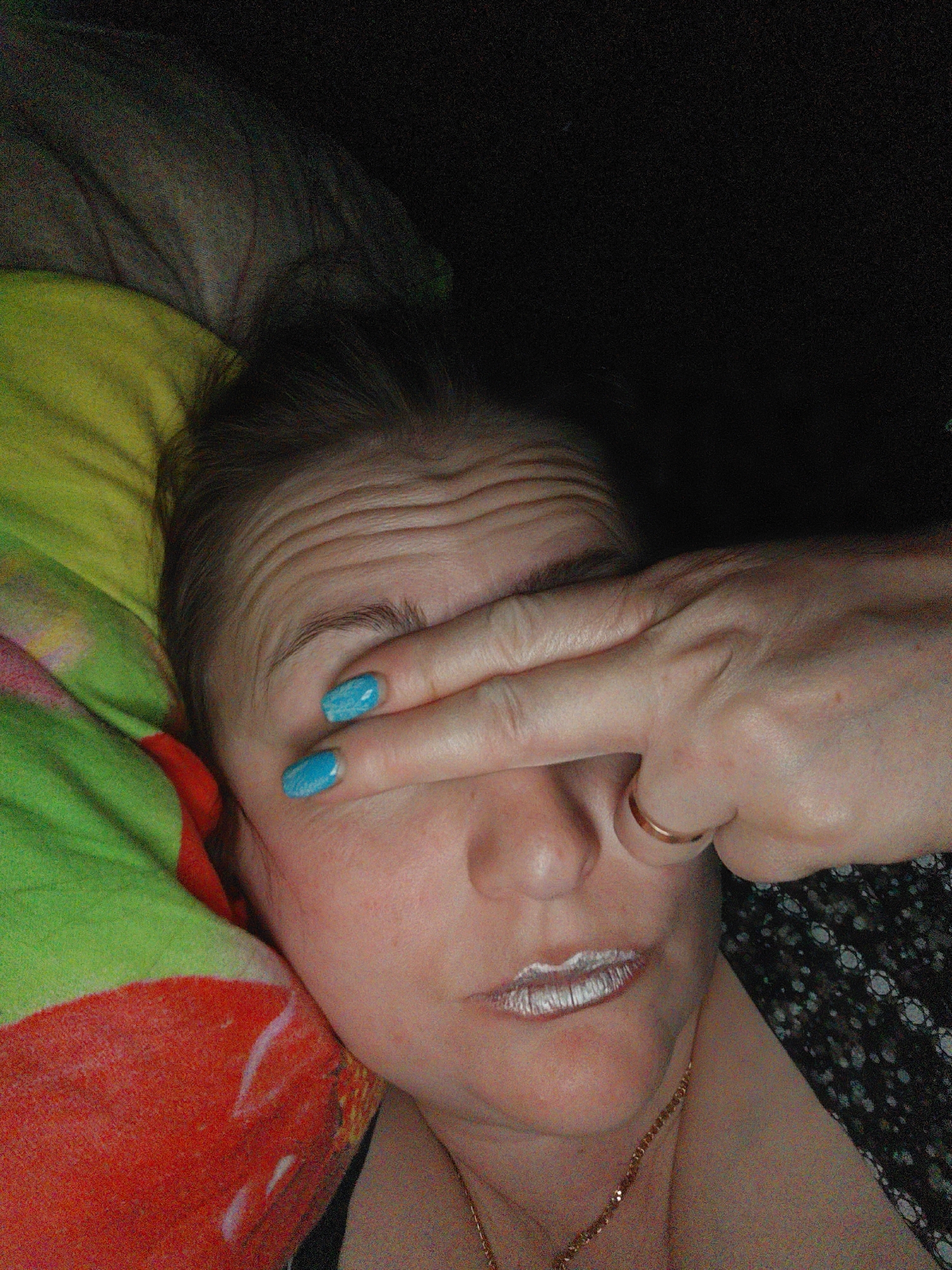 A person lies on their back with a patterned pillow partially visible on the left side of the frame. They are covering their eyes with their right hand. The person has blue nail polish and wears a gold ring on their ring finger. Their lips are painted silver. A gold necklace is visible around their neck. The background is dark and largely out of focus. There is no specific evidence in the image to confirm a location in the city of Фоки, Russia.