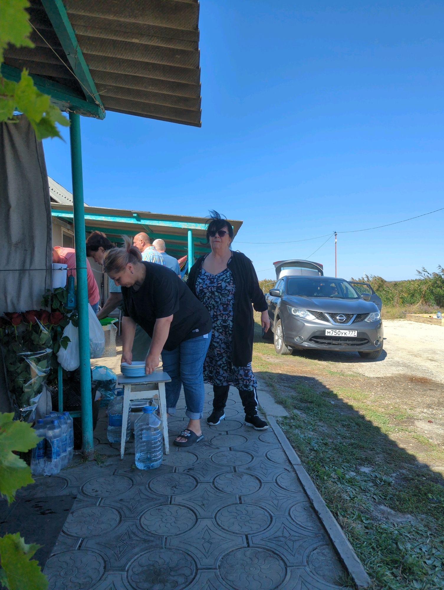 In Прикумское, Russia, a group of people are gathered outdoors near a grey Nissan Qashqai with an open trunk. A woman in a black t-shirt and blue jeans is stacking blue and white plates on a small stool. Another woman in a patterned dress and black cardigan stands to her right, looking towards the camera. Several other individuals are visible in the background, some near what appears to be a food serving area. Stacks of bottled water and a bouquet of red roses are on the left side of the image. The ground is paved with patterned tiles, and a grassy area is visible with dry vegetation in the distance under a clear blue sky.