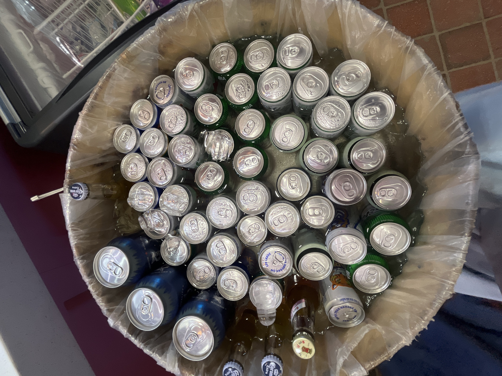 A top-down, close-up shot shows a clear plastic-lined bin filled with an assortment of canned beverages and bottles submerged in water, likely for chilling. The bin is situated on a brick-tiled floor. Among the visible items are numerous silver aluminum cans with various labels, some green cans, and several brown glass bottles with distinct yellow and white caps, one of which is identifiable as "Corona Extra." A few cans appear opened or partially submerged. The background reveals a partial view of a refrigeration unit, suggesting the bin might be located in a commercial establishment, possibly a bar or convenience store in Đà Nẵng, Vietnam, given the context of the "Corona Extra" branding. The overall scene suggests a casual setting where beverages are being kept cool.