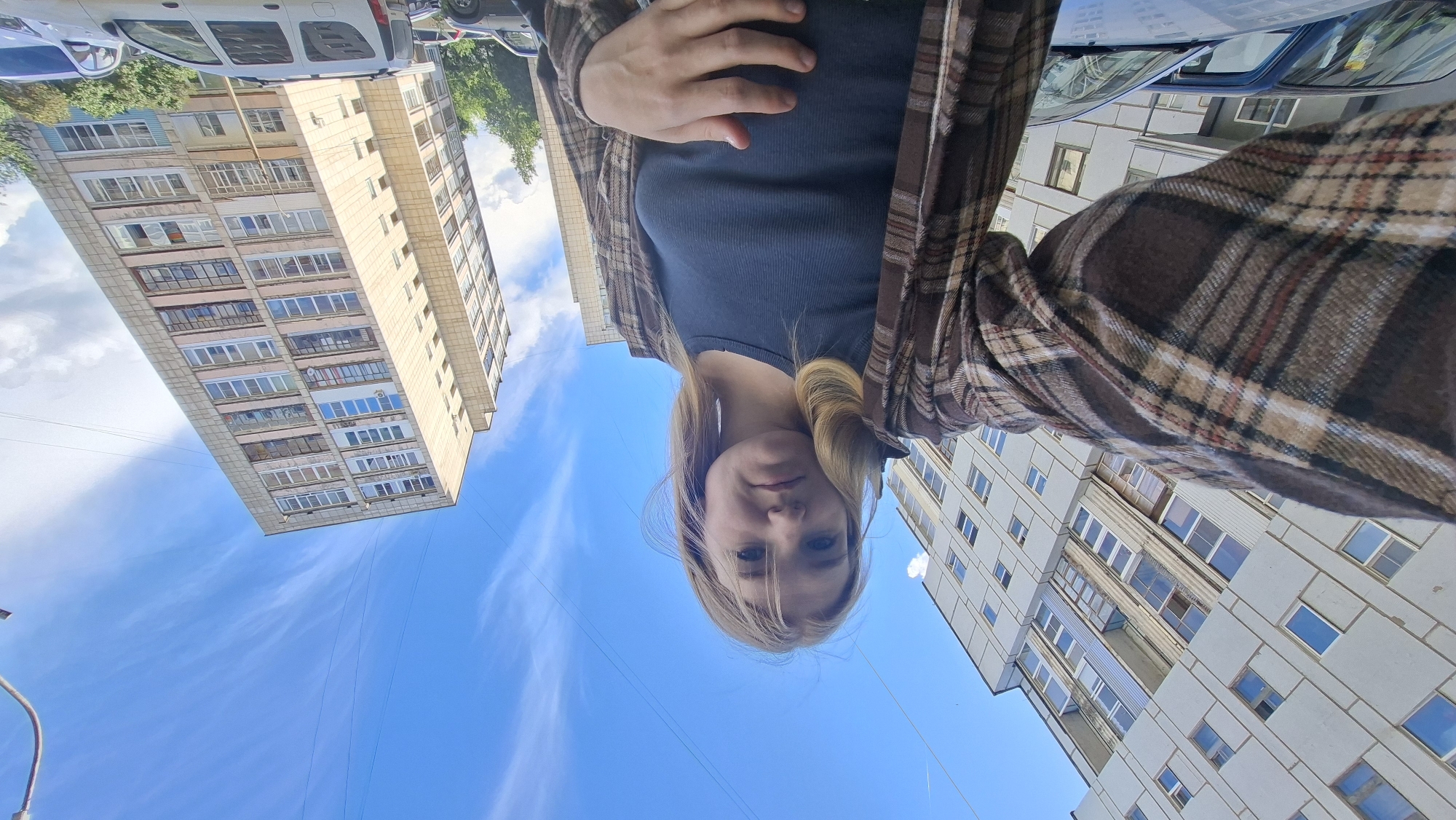 A young woman with blonde hair is taking a selfie in an outdoor urban environment. She is wearing a brown and tan plaid shirt over a dark ribbed tank top, and her left hand is resting on her abdomen. Behind her, tall, multi-story apartment buildings with many windows are visible against a bright blue sky with wispy white clouds. Cars are parked in front of the buildings. The architecture suggests a typical residential area in a Russian city like Chaikovsky.