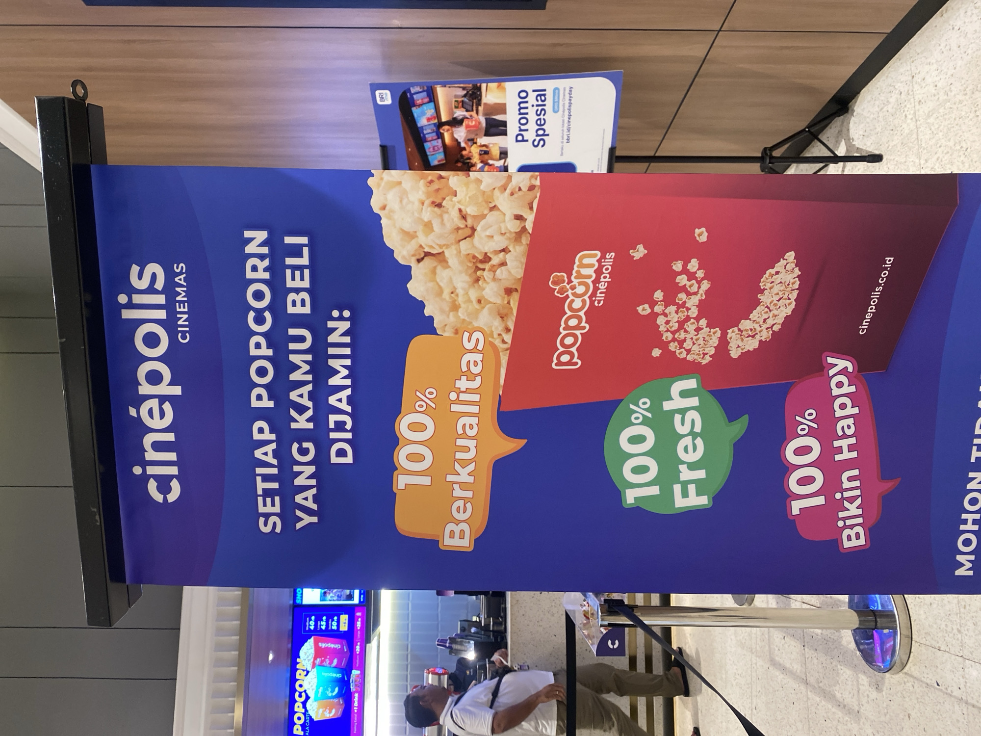 The image shows a vertical banner for Cinépolis cinemas in Banjarbaru, Indonesia, advertising popcorn. The banner is predominantly blue and features text in white and orange. The top of the banner displays the Cinépolis logo and the word "CINEMAS". Below this, the text "SETIAP POPCORN YANG KAMU BELI DIJAMIN:" is prominently displayed, meaning "Every popcorn you buy is guaranteed:".
The banner highlights three key features of the popcorn: "100% Berkualitas" (100% Quality) in an orange speech bubble, "100% Fresh" in a green speech bubble, and "100% Bikin Happy" (100% Makes Happy) in a pink speech bubble. An illustration of a red popcorn box filled with popcorn is on the right side of the banner, with a scattering of popcorn kernels below it. The website "cinepolis.co.id" is also visible at the bottom of the banner.
To the left of the main banner, a digital screen shows prices for popcorn and drinks, with options like "1 Drink" and different popcorn sizes. A man in a white shirt and khaki pants is standing near a counter, appearing to be a customer or employee. Metal stanchions with dark ropes are present, indicating a queueing area.
A second, smaller banner is partially visible to the right of the main banner. This banner has a white background and features text in blue and orange. It advertises "Promo Spesial" (Special Promo) and includes a QR code and a website address. The BRI (Bank Rakyat Indonesia) logo is visible at the top of this smaller banner, suggesting a partnership.
The overall environment suggests a cinema lobby or concession area, with tiled flooring and wooden-paneled walls in the background. The lighting is bright, typical of an indoor public space. The location is specified as Banjarbaru, Indonesia.