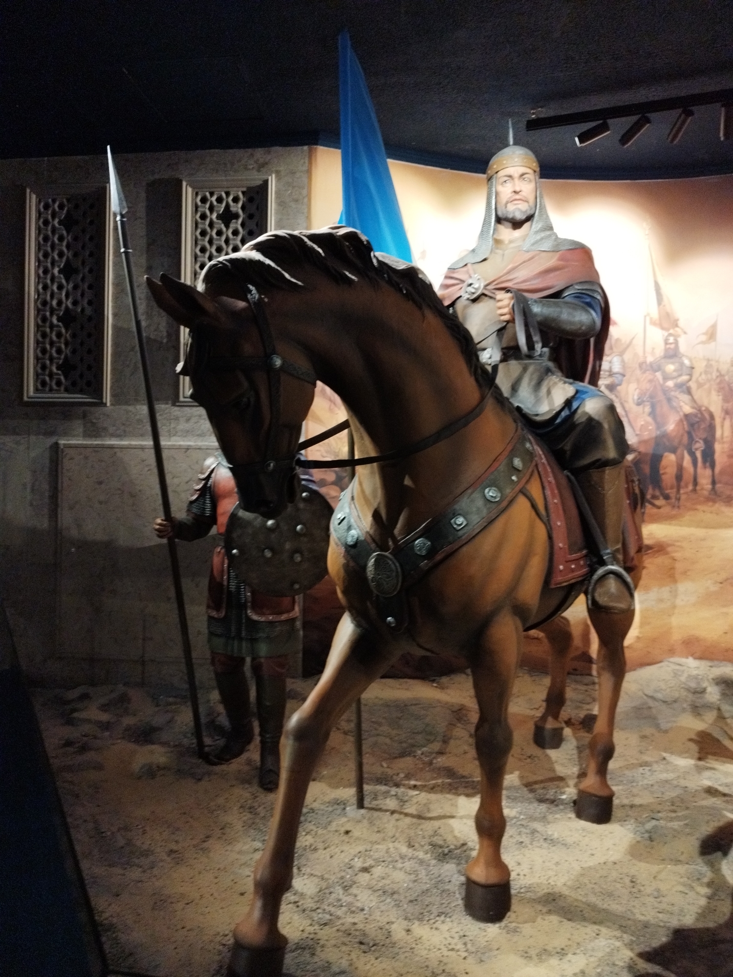 A diorama depicts a mounted warrior and a foot soldier in a staged historical setting. The mounted warrior, a man with a beard and wearing a helmet, chainmail, and a red cloak, is seated on a brown horse. He holds a spear in his left hand. To the left of the horse, a foot soldier in armor and a red tunic stands holding a spear. The background features a mural of a battle scene with more mounted soldiers. The diorama is displayed indoors, with dark walls and ceiling, and lighting from above. The floor is covered in sand and gravel. The overall presentation suggests a museum or historical exhibit. The location is listed as Unknown, Unknown.