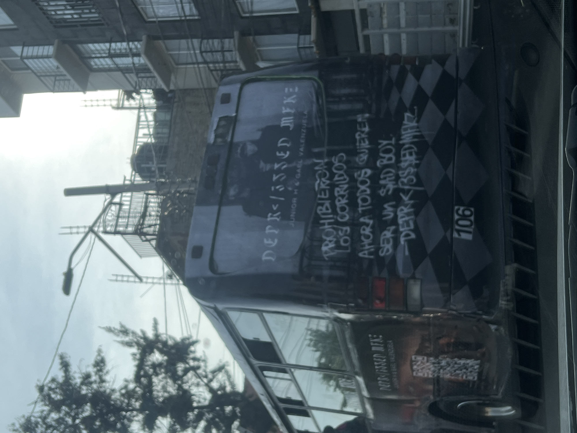 The image shows the back of a bus in Mexico City, Mexico. The bus is gray and has a checkerboard pattern painted on the back. There is graffiti written on the back of the bus that includes the words "DEPR<$SED MFKE," "JUNIOR H & GAEL VALENZUELA," "PROHIBIERON LOS CORRIDOS," "AHORA TODOS QUIEREN SER UN SAD BOY," and "DEPR</3$$ED MFKZ." The bus also has the number "106" painted on the back. The bus is parked on a street in Mexico City, and there are buildings and trees visible in the background.