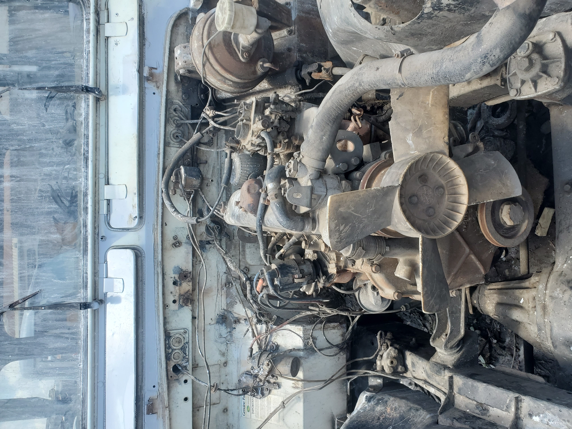 The image shows the engine compartment of an old, weathered vehicle, likely a truck or bus, in Rafin Par, Nigeria. The engine is dirty and appears to be in a state of disrepair, with many wires exposed and parts covered in dust and grime. The overall impression is one of neglect and abandonment. The environment is unkempt, with debris visible on the ground. There are no people present in the image.