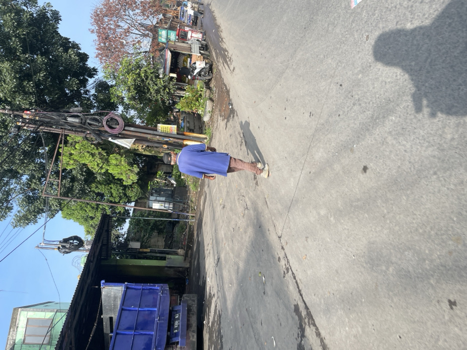 The image shows a man walking down a street in Bandung, Indonesia, at the location -6.927, 107.584. He is wearing a blue shirt and brown pants. The street is paved with concrete and has a few small cracks. The man's shadow is cast in front of him. There is a truck parked on the left side of the street and some trees and vegetation on the right side. The scene is a mix of residential and commercial areas. The weather appears to be sunny and clear. There are several power lines and poles in the background. The man is walking toward the right of the image.