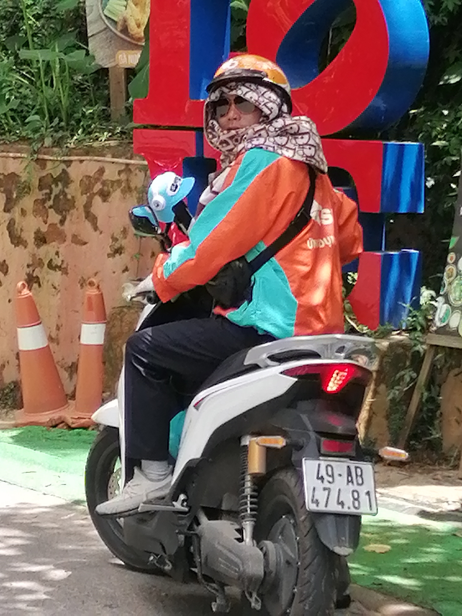 A person wearing a helmet, sunglasses, and a patterned scarf over their face is riding a white scooter in Da Lat, Vietnam. The rider is also wearing an orange and teal jacket and dark pants. The scooter has a license plate reading "49-AB 474.81". In the background, there are large red and blue block letters, two orange traffic cones, and lush green foliage. The ground appears to be paved with some areas of green artificial turf. The overall scene suggests a person on a scooter in an urban or tourist area.