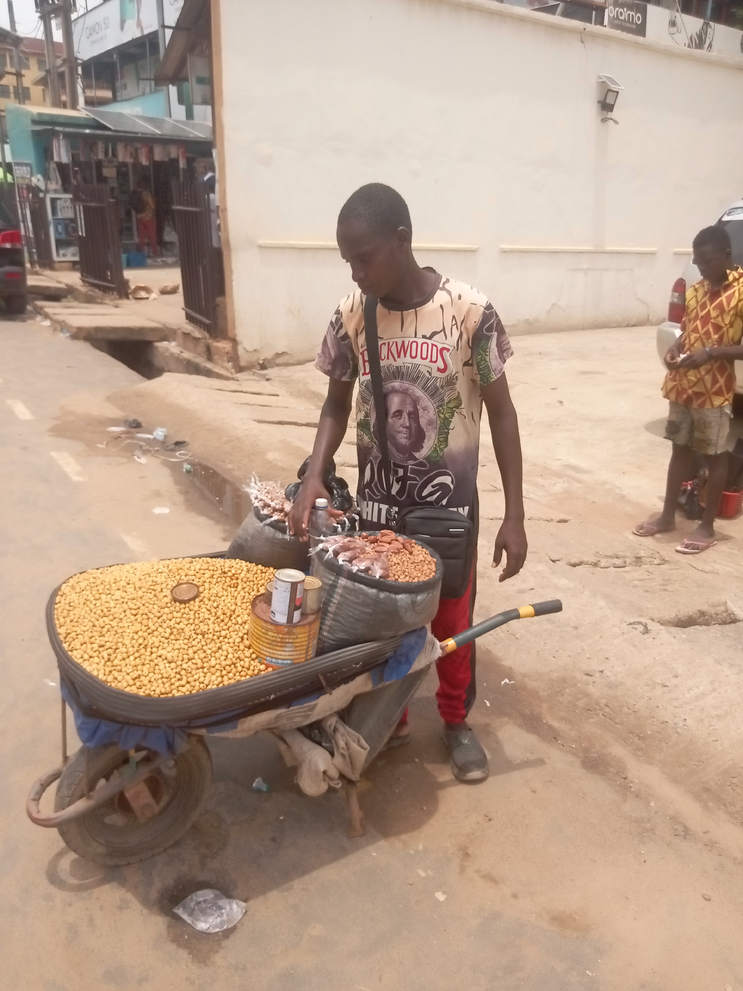 In Fegge, Nigeria, a street vendor is attending to a wheelbarrow filled with goods for sale. The wheelbarrow contains a large quantity of round, yellow seeds, likely nuts or grains, along with smaller containers and bags of other items. The vendor is a young man wearing a t-shirt and red pants, with a shoulder bag. Another person stands nearby. The setting is an urban street with buildings and evidence of commercial activity in the background.