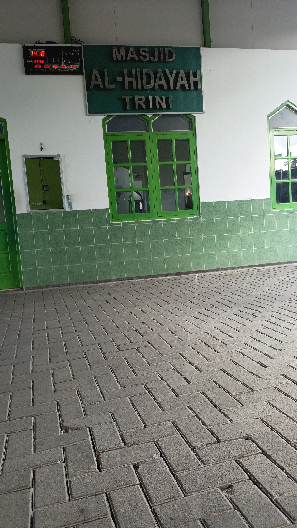 An exterior view of the AL-HIDAYAH mosque in Trihanggo, Indonesia, shows a section of its facade and the paved area in front. A digital clock displaying "04:18" is mounted on the wall, above a sign that reads "MASJID AL-HIDAYAH TRINI". A green-framed window with multiple panes is prominent, and another smaller green-framed window is visible to the right. The lower portion of the building is covered in green square tiles. The ground in front of the mosque is paved with grey rectangular interlocking bricks laid in a herringbone pattern. The scene is illuminated by natural light, casting shadows.