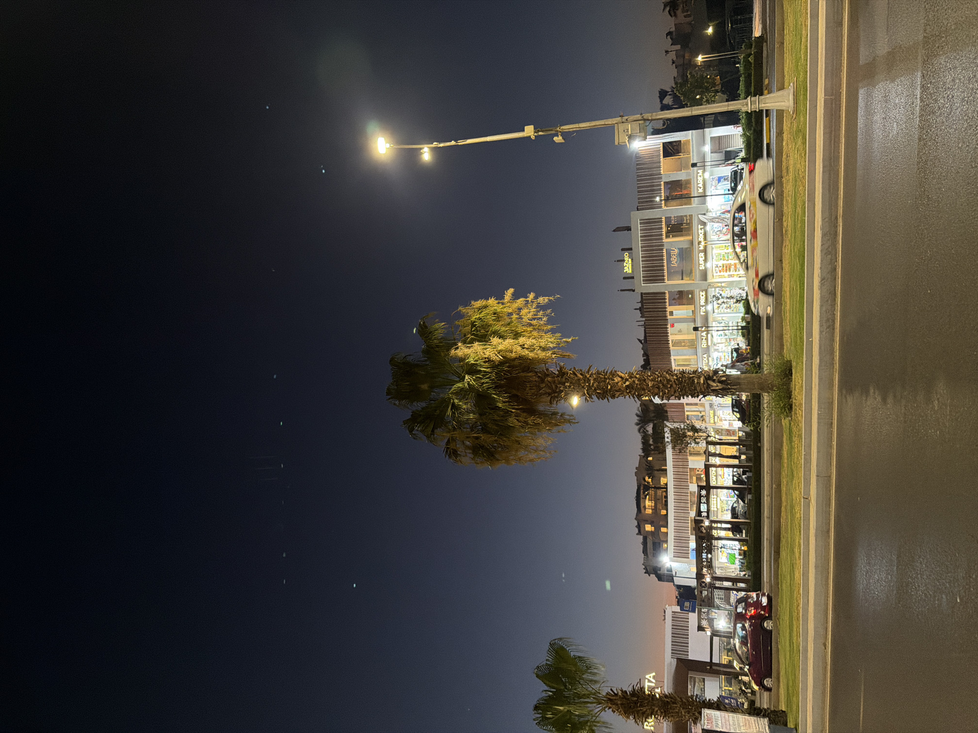 The image depicts a nighttime scene on a street in Sharm El Sheikh, Egypt. A tall street lamp illuminates the area with warm light. In the foreground, a palm tree with green fronds stands prominently. To the right, a modern building with multiple levels houses shops and businesses, indicated by illuminated windows and signage.
Visible text on the building includes "KAREM", "JAZZCO", "SUPER MARKET", "FIX PRICE", "RIHA", "ANTHEKA", and "RIHA". The storefronts suggest a commercial area. A white car with colorful decals is parked in front of one of the shops, and a red car is visible further down the street. The street itself appears wet, suggesting recent rain or mist. The sky is dark blue, with a few faint lights or stars visible. The overall ambiance is that of a lively commercial street at night, likely a tourist area given the location context. The lighting creates a somewhat atmospheric and inviting scene.