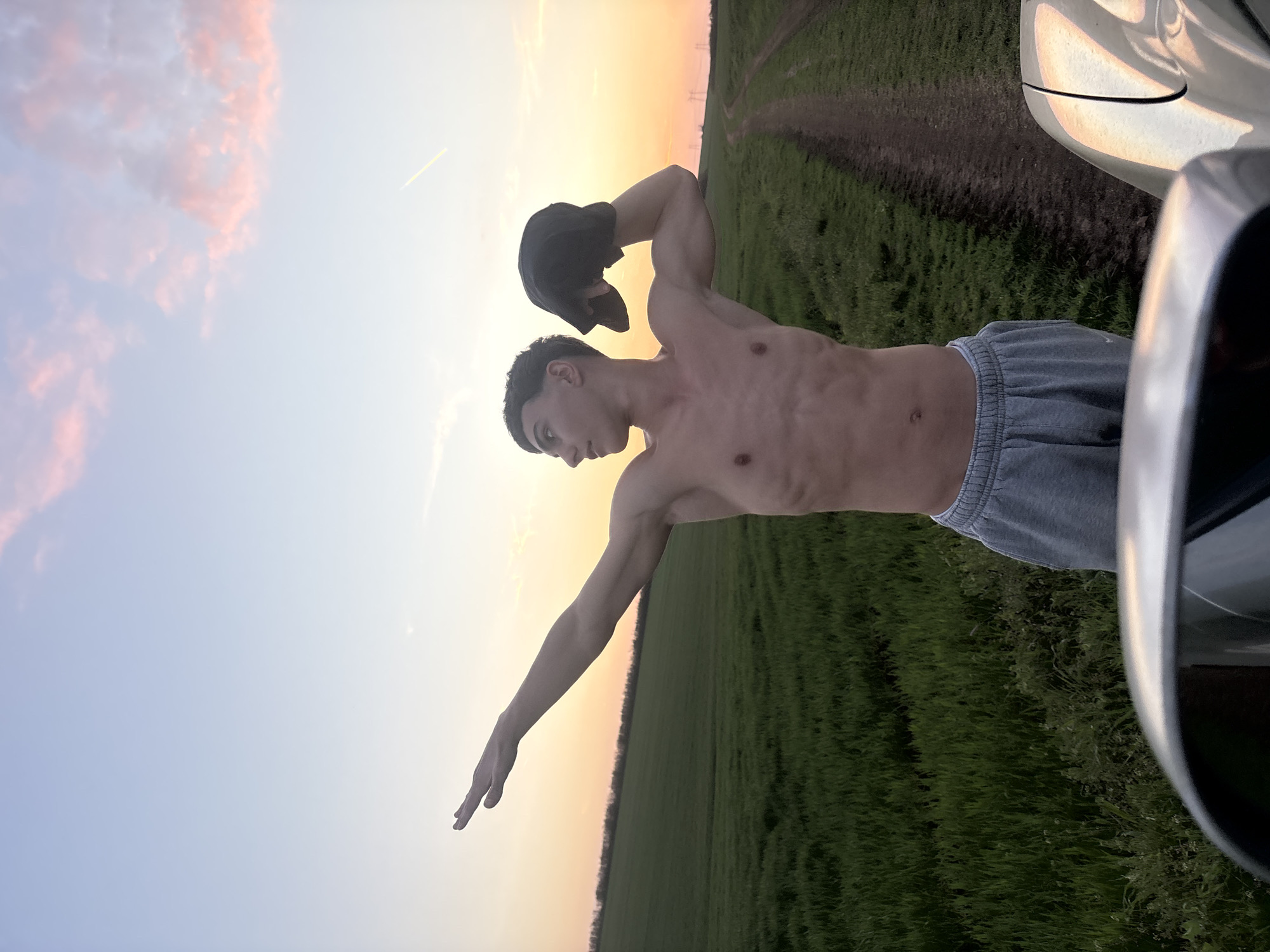 A shirtless male stands in a field, holding a black hat over his bicep. He wears gray shorts. A car is partially visible to the right. The background is a field and a sunset sky. This scene is located in Lehchevo, Bulgaria.