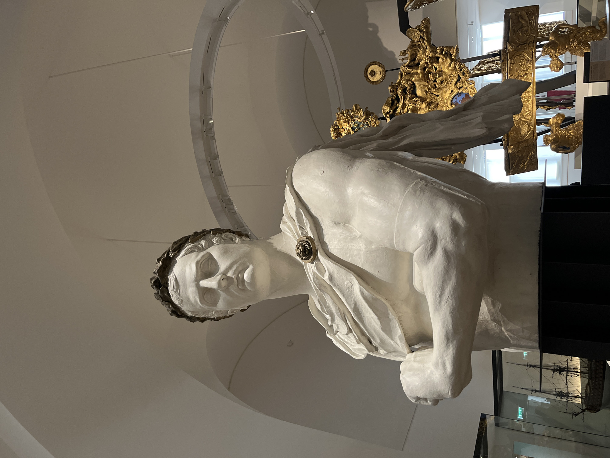 A marble bust of a man with a laurel wreath is displayed in a museum. The bust depicts a stern-faced individual with arms crossed and a draped cloth over his shoulder, fastened with a circular emblem. The bust is placed on a black pedestal. Behind the bust, to the right, a golden ornate clock or timepiece is visible, featuring intricate carvings and figures. Further to the left, a model ship in a glass display case can be seen. The environment suggests an indoor exhibition space with a high, arched ceiling. The lighting is soft and ambient. The specific location within Paris, France, is not determinable from the image itself.