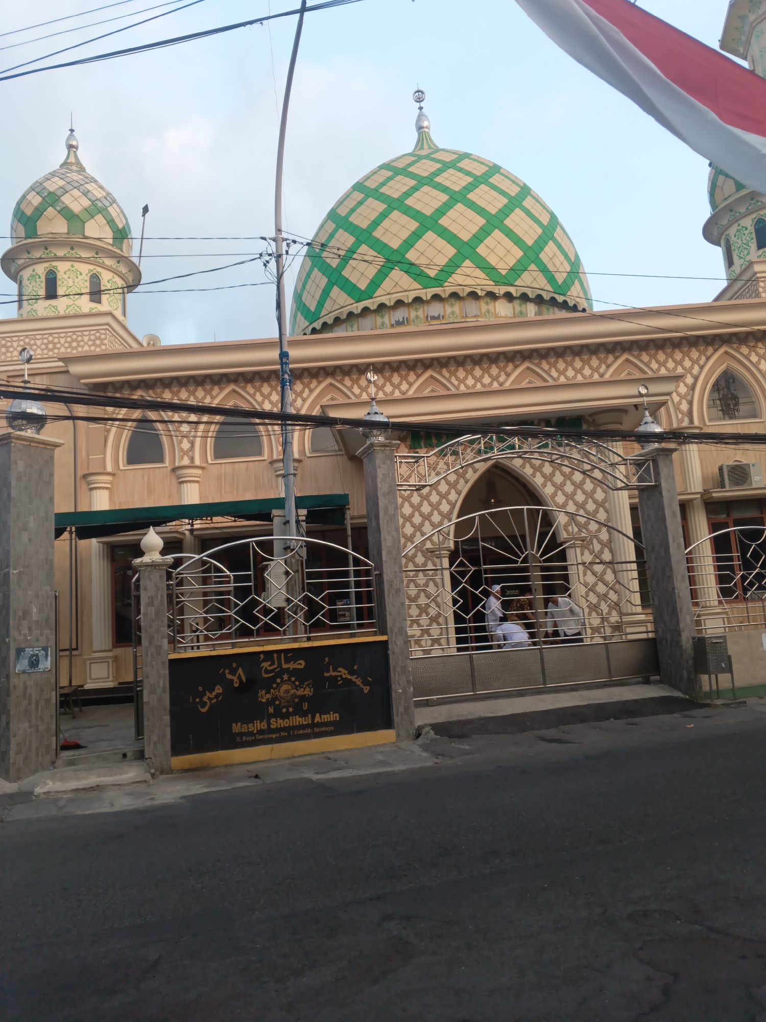 The image shows the facade of the Masjid Sholluhul Amin in Surabaya, Indonesia, located at Jl. Raya Semampir No. 1 Sukolilo. The mosque features two prominent domes, one larger than the other, both adorned with a green diamond pattern. The building's exterior is a warm beige color with intricate decorative elements around the windows and along the roofline. A metal gate, decorated with geometric patterns, stands in front of the mosque's entrance. Several people, dressed in white, are visible entering or standing near the entrance. Overhead, electrical wires crisscross the sky, and a portion of an Indonesian flag is visible in the upper right corner. The foreground shows a street with asphalt paving.
