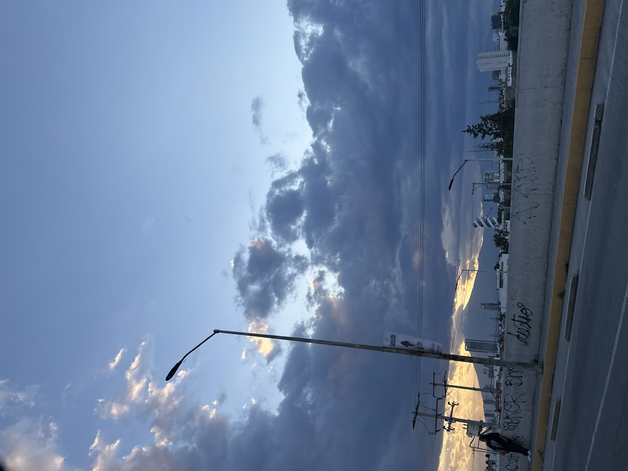 The image shows a cityscape under a dramatic sky with dark, heavy clouds interspersed with patches of blue. A concrete barrier runs along the right edge of the frame, indicating a bridge or elevated road. Graffiti adorns the barrier. On the left, a street lamp is visible, with power lines extending from it. In the background, buildings of varying heights make up the city skyline. A person is standing near the base of a power pole, next to the barrier. The location is Unknown, Unknown.