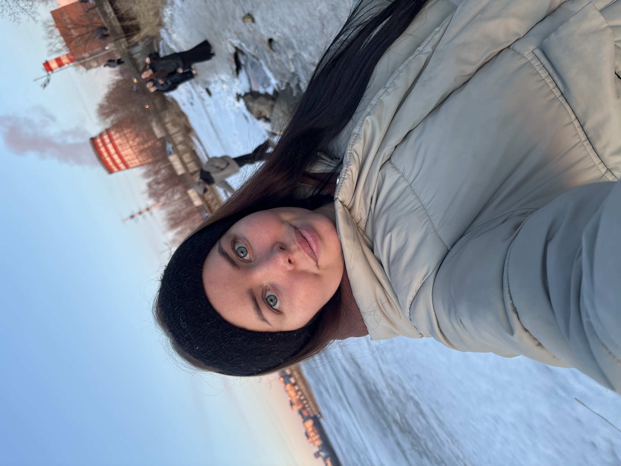 A woman takes a selfie outdoors, wearing a black knit cap and a light beige puffer jacket. Her hair is long and dark. In the background, there is a partially frozen body of water with what appears to be a building with red and white stripes, possibly a factory, emitting some smoke. There are other figures discernible in the background near the water's edge. The scene suggests a winter setting with the sky a pale blue at the horizon.
Given the visual cues, the location is likely near the Izhevsk Pond in Izhevsk, Russia, where a similar factory building (Izhevsk CHP-1) exists along the shore.