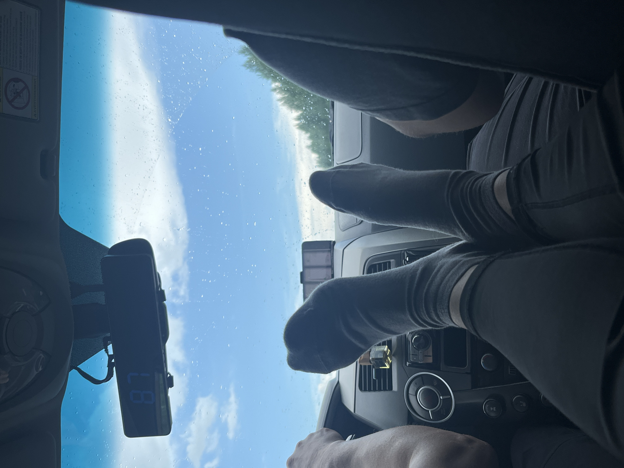 The image shows the interior of a vehicle, likely a van or SUV, from the perspective of the back seat passengers. The focus is on the feet of two individuals wearing dark gray socks, extended forward towards the dashboard. One person's left arm is visible, resting on the steering wheel. The dashboard is visible, with a digital clock displaying "87" mounted on the rearview mirror. The windshield shows a cloudy sky with raindrops, indicating it is raining. Outside, a line of trees can be seen. The vehicle appears to be in motion. The location is specified as Goldyrevskii, Russia.
