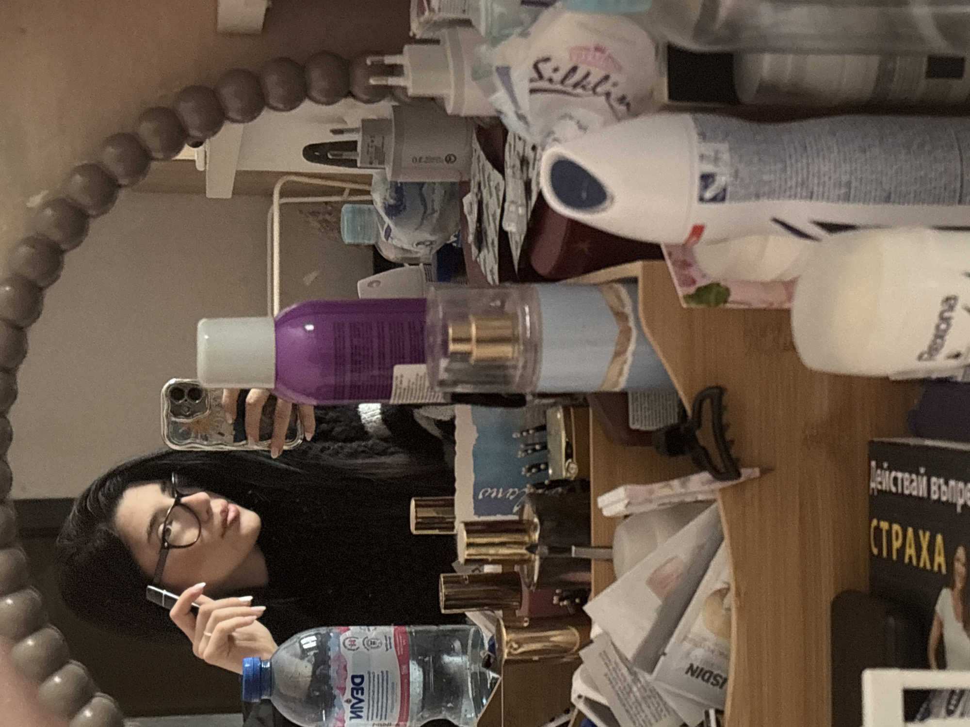 A young woman with black hair and glasses is taking a selfie in a mirror. She is holding a phone in one hand and a black object, possibly makeup, in the other. The mirror has a beaded frame. The background is cluttered with various toiletries, cosmetics, and other items. There is a bottle of DEVIN water visible. A book with writing in Cyrillic is also present. The image could be taken in Sofia, Bulgaria, given the Cyrillic text on the book.