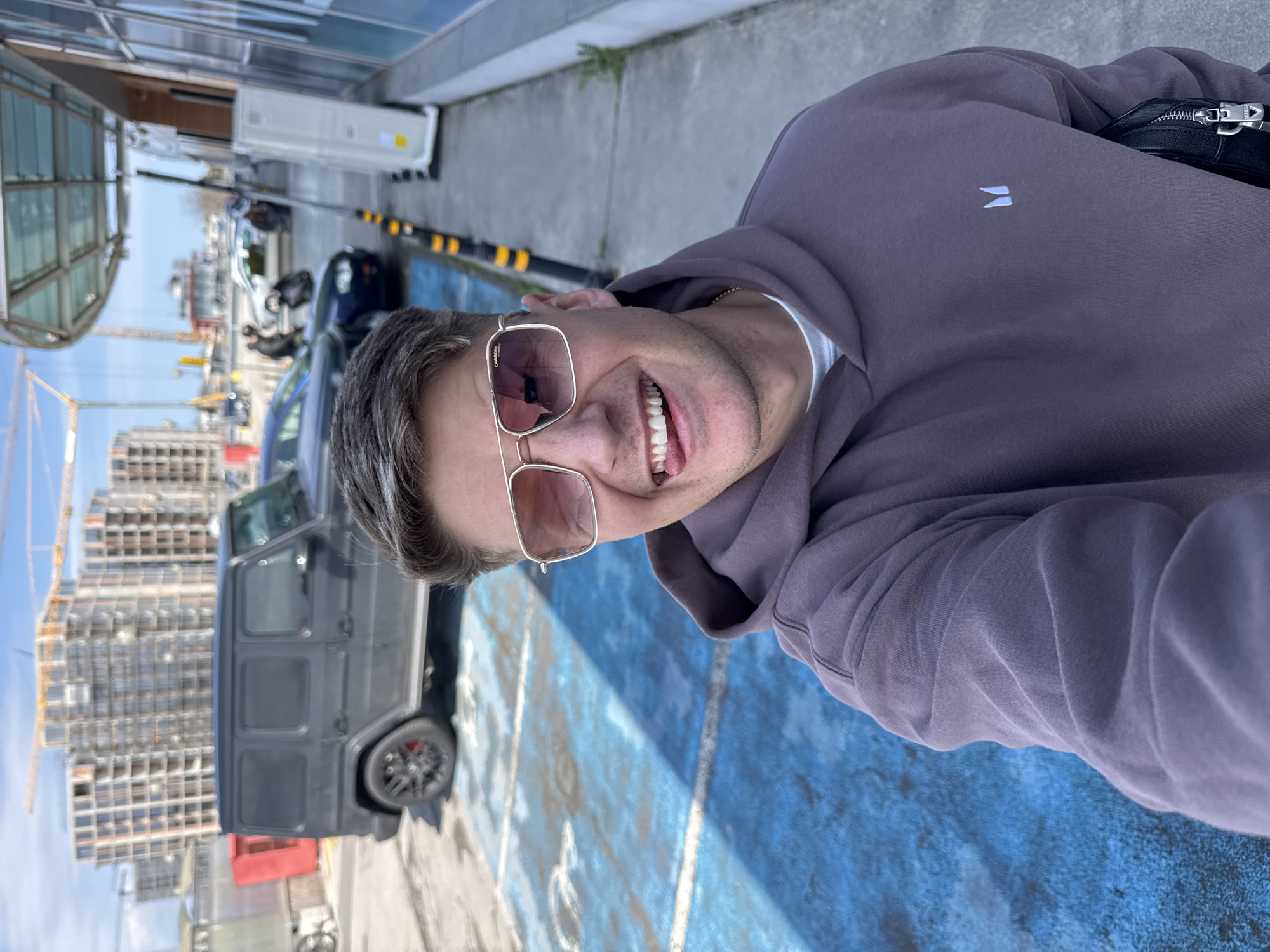 A man is taking a selfie outdoors in what appears to be a parking lot or urban area. The man is wearing sunglasses, a purple hoodie, and a white undershirt. He is smiling and looking directly at the camera.
Behind the man is a black Mercedes-Benz G-Wagon parked in a blue-painted handicap parking space. Further in the background, construction cranes and buildings are visible, indicating ongoing construction activity in the area. Based on the presence of a Mercedes-Benz G-Wagon, construction cranes, and the architecture, it is likely the location is in a more affluent area within Burgas, Bulgaria, such as near the port or central areas undergoing development. Other parked vehicles are also visible in the background. The sky is blue.