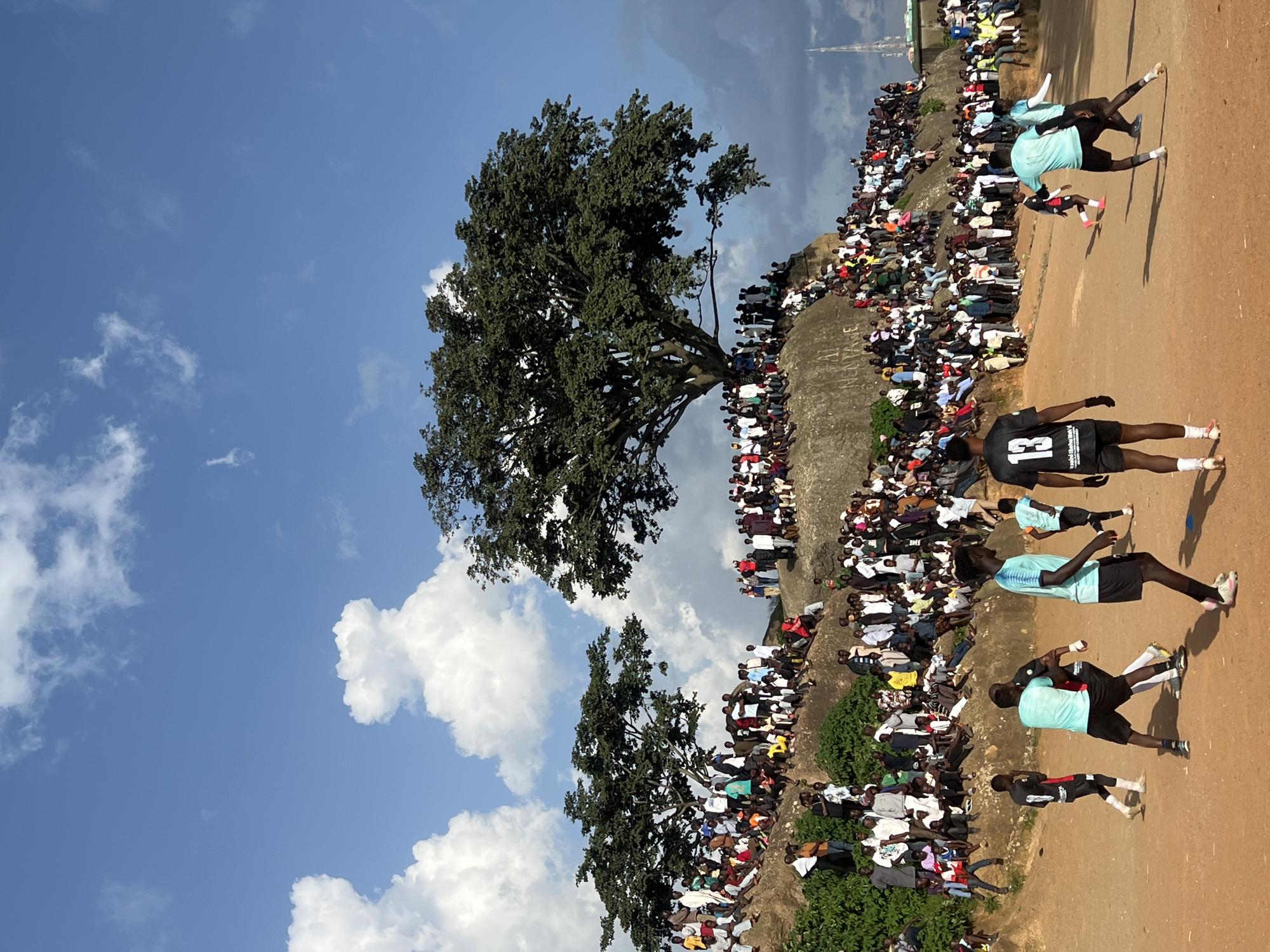 The image depicts an outdoor gathering of people in Jos, Nigeria, specifically at the popular tourist site known as "Jos Rock" or "Assop Falls." A large crowd is densely packed on a rocky hillside, with many individuals seated or standing to observe an event unfolding on the dusty ground below. Two prominent trees with full, green canopies dominate the background, casting shadows on the spectators. The sky is a clear blue with scattered white clouds.
In the foreground, several young men are engaged in what appears to be a soccer match. One player, wearing a black jersey with the number 13, is positioned facing away from the camera. Another player in a turquoise jersey is actively participating in the game, with a third player in a similar turquoise jersey moving past him. A fourth player, in a black and white jersey, is visible further in the background, also engaged in the game. The ground is a reddish-brown earth, typical of the Jos Plateau. In the far distance, a tall, slender structure, possibly a communication tower, can be seen. The overall scene suggests a vibrant community event taking place in a natural, open-air setting.