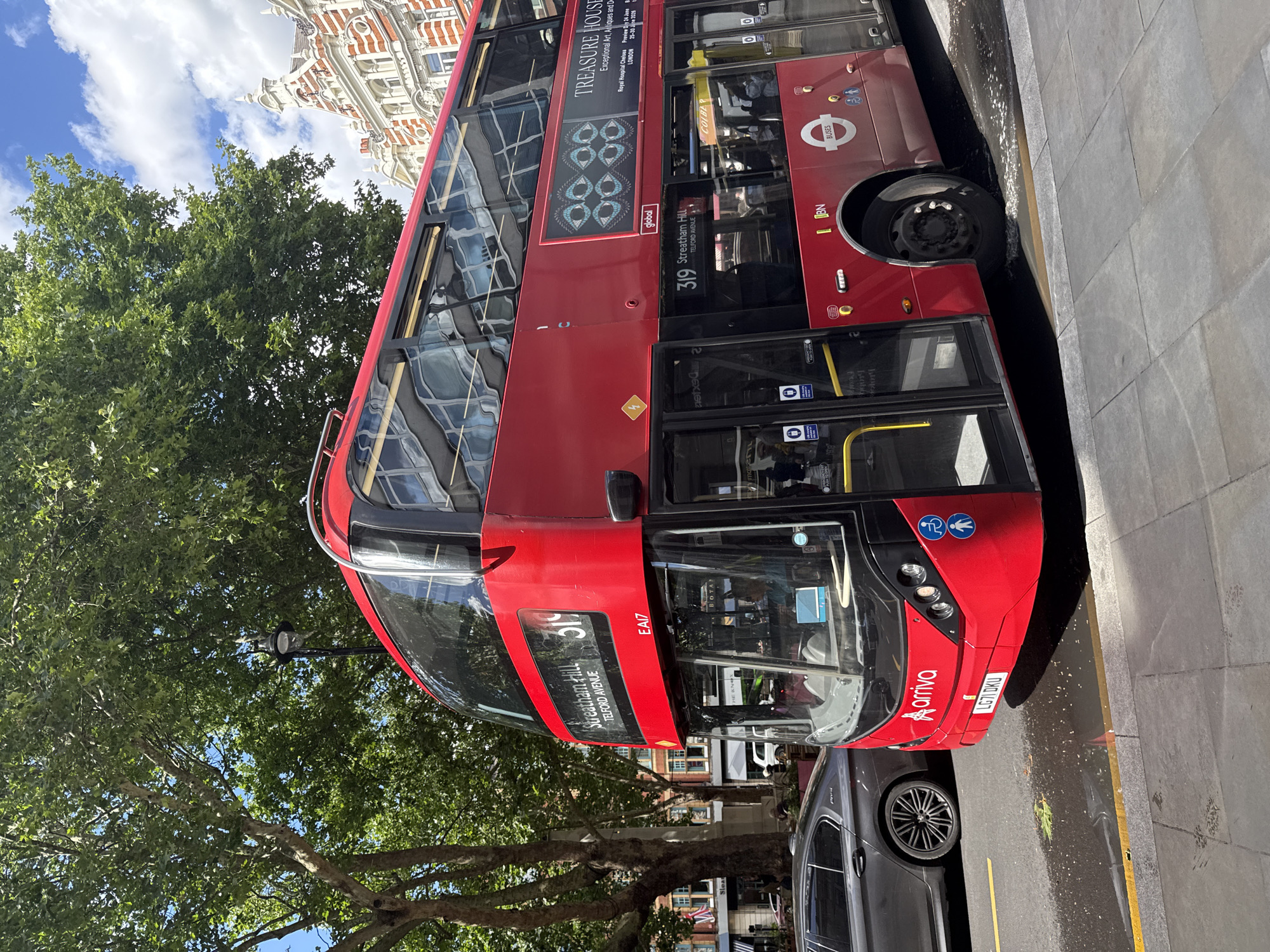 A bright red, double-decker Arriva bus, route 319 to Streatham Hill via Telford Avenue, is prominently featured, angled towards the viewer from the right side of the frame. The bus has the license plate LG71 Dku. On its side, an advertisement displays "TREASURE HOUSE" with an artistic representation of eyes above the text.
The bus is on a city street, with a grey car parked to its left. The road has yellow and white lines marking the lanes. In the background, a grand building with white and red ornate architecture stands, suggesting a well-established part of London. Lush green trees with full canopies overhang the street, and the sky is a clear blue with scattered white clouds. A lamppost with a distinctive design is visible between the trees and the bus. The bus is in motion, as indicated by the slight blur on the wheels. The scene depicts a typical urban environment in London on a sunny day.