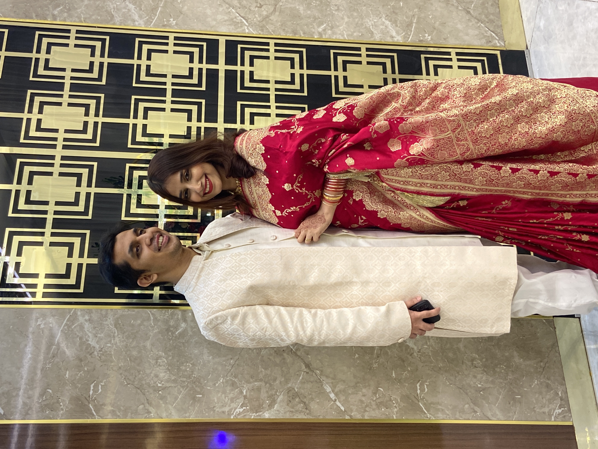 In Dhaka, Bangladesh, a man and a woman pose for a photograph indoors. The man lies horizontally, wearing a light beige sherwani over white pants, holding a black object in his right hand. The woman lies atop him, draped in a red and gold saree. She has bangles on her right wrist and henna on her hand. Both are smiling and looking towards the camera. Behind them is a decorative wall panel with a black and gold geometric design. The floor is covered with light brown marble tiles with brown trim. A blue light is visible at the bottom left of the frame.