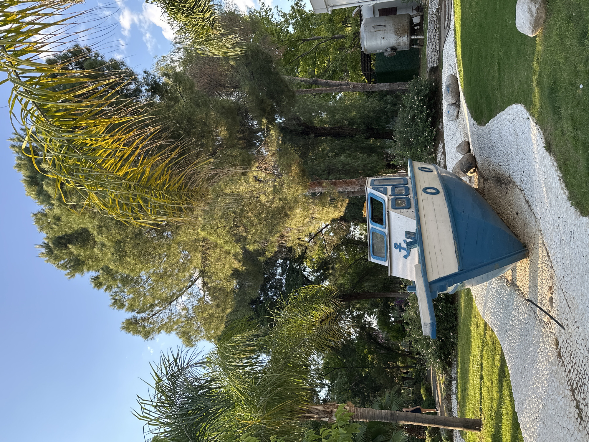 The image shows an outdoor scene in Fethiye, Turkey, featuring a blue and white boat resting on a bed of white pebbles next to a green lawn. The boat's cabin is visible, and an anchor emblem is displayed. Tall trees, including palm trees, surround the boat. In the background, a structure resembling a water tank is visible. The sky is blue with scattered clouds.