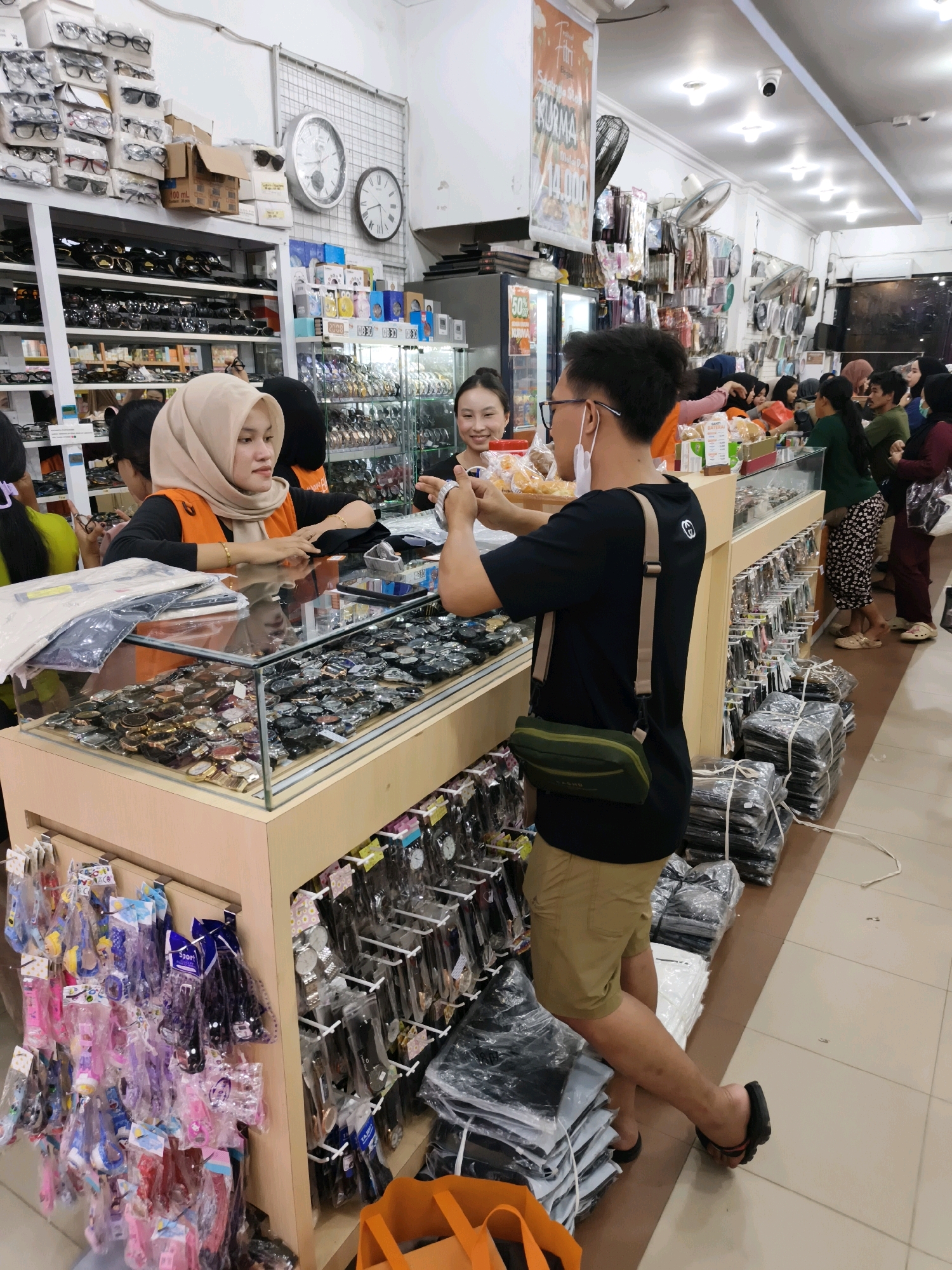 In a retail establishment in Ketapang, Indonesia, a customer, a man wearing glasses, a black t-shirt, and tan shorts, is looking at items on display at a counter. He is holding a product with both hands and appears to be examining it. To his left, a female shop employee wearing a hijab and an orange vest is interacting with the customer. Behind the customer and employee, other shoppers and employees are visible browsing and working. The counter displays numerous watches and other accessories. The store itself appears to be well-stocked, with shelves filled with various goods like eyeglasses and more watches. The lighting suggests it is daytime, likely indoors.