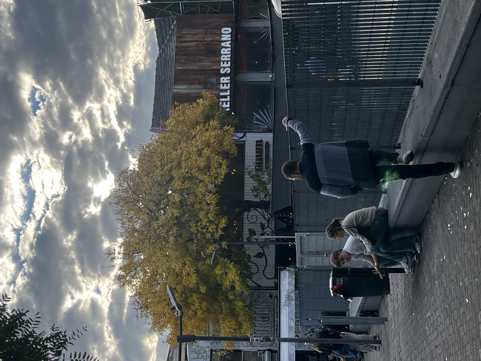 The image is a street scene in Buenos Aires, Argentina, on a cloudy day. A building is labeled "KELLER SERRANO." A large tree with yellow leaves stands near the building. Several people are present. One man is standing near a metal fence, with his arm raised. Two people, one wearing a white shirt and the other a light sweater, are positioned near a trash can on the sidewalk. Further down the street, there are additional people visible. The ground is paved with bricks.