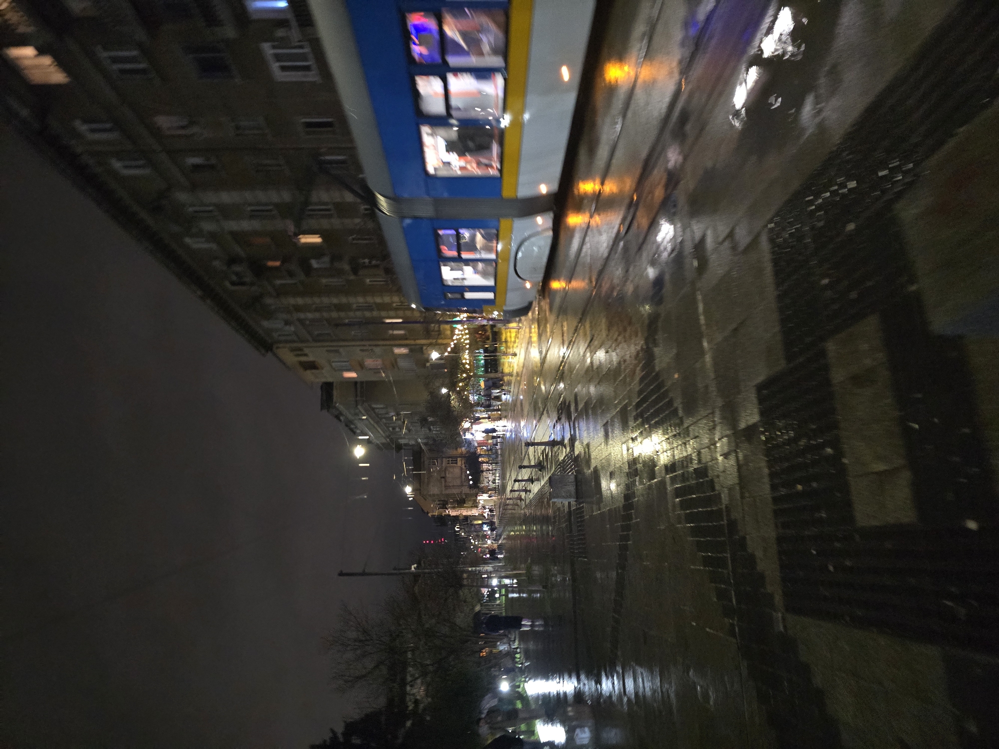 The image shows a street scene in Sofia, Bulgaria at night, after a rain shower. The street is wet and reflective, with reflections of streetlights and buildings. A tram is passing by on the left side of the street. The sidewalk has a distinctive patterned design. Buildings line the left side of the street, with lit windows. In the distance, people are walking along the street. The sky is dark, and there are trees and vegetation visible.