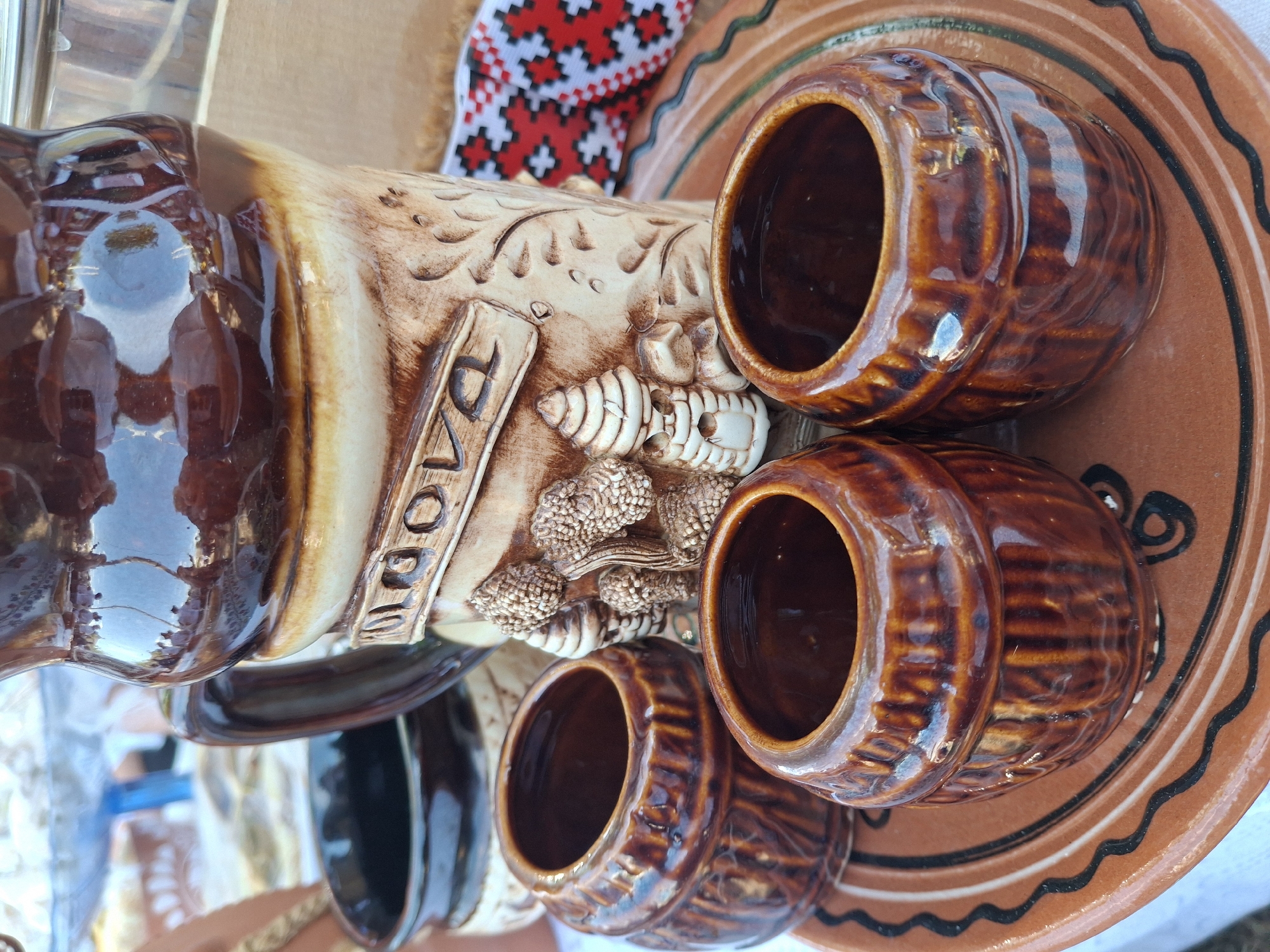 A close-up, eye-level shot captures a rustic ceramic set on a table, likely in a market or outdoor setting in Roghi, Moldova. The primary subject is a large, brown, glazed pitcher with a cream-colored band featuring embossed details of leaves and what appear to be small, tiered structures resembling hives or decorative elements. The word "MOOLA" is clearly visible, embossed in a rectangular frame on the cream band. The pitcher's surface reflects a brightly lit outdoor environment with hints of people and structures.
In front of the pitcher, three smaller, barrel-shaped ceramic cups, also with a dark brown glaze and ribbed detailing, are arranged on a larger, circular, terracotta-colored ceramic plate. This plate has decorative black and white wavy lines around its rim. The overall impression is one of traditional craftsmanship. The background is softly blurred, showing more of the table setting, including a dark-colored ceramic vessel on the left and a piece of fabric with a red, black, and white geometric pattern on the right. The lighting suggests it is daytime.