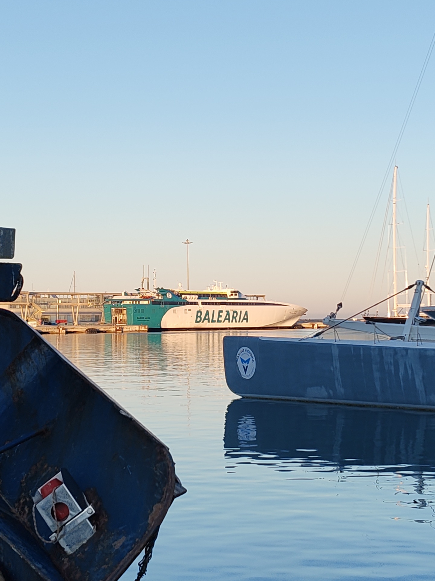 The image shows a harbor scene in Dénia, Spain, at dusk or dawn, evidenced by the soft, diffused light. In the background, a large, white passenger ferry with the name "BALEARIA" prominently displayed is docked at a pier. The ferry features green accents and a modern design. Midground shows one yacht and the masts of others and the foreground presents sections of a two smaller boats. The water is still and reflects the colors of the sky and the surrounding boats, contributing to the overall calm atmosphere of the harbor. The scene lacks people.