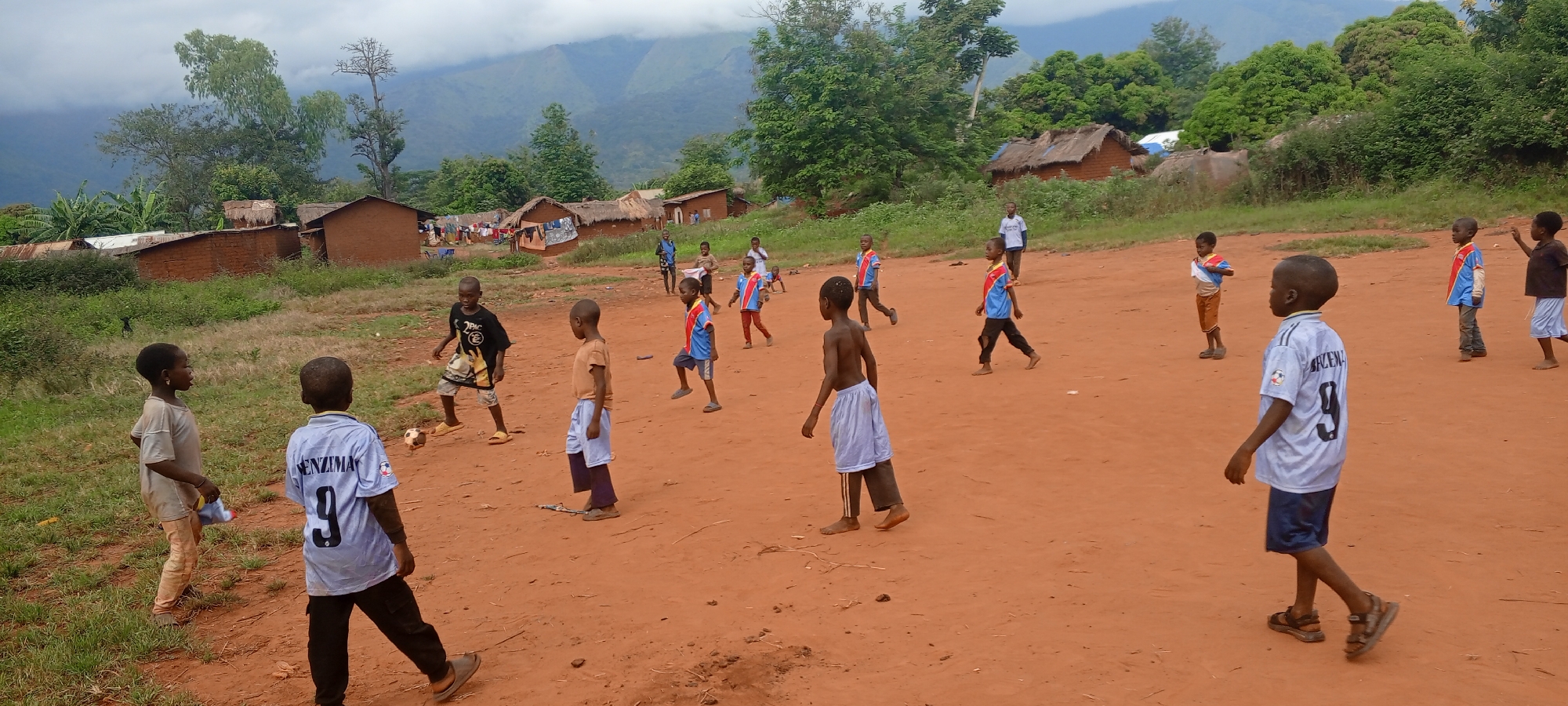Children are playing soccer on a dirt field in Ngalula, Democratic Republic of the Congo. Approximately 15 boys are visible, some in soccer jerseys, others in casual clothing. In the background are small brick houses with thatched roofs, surrounded by green vegetation and mountains obscured by clouds. One adult male is standing near the houses. The ground is red clay, typical of the region. The sky is overcast.