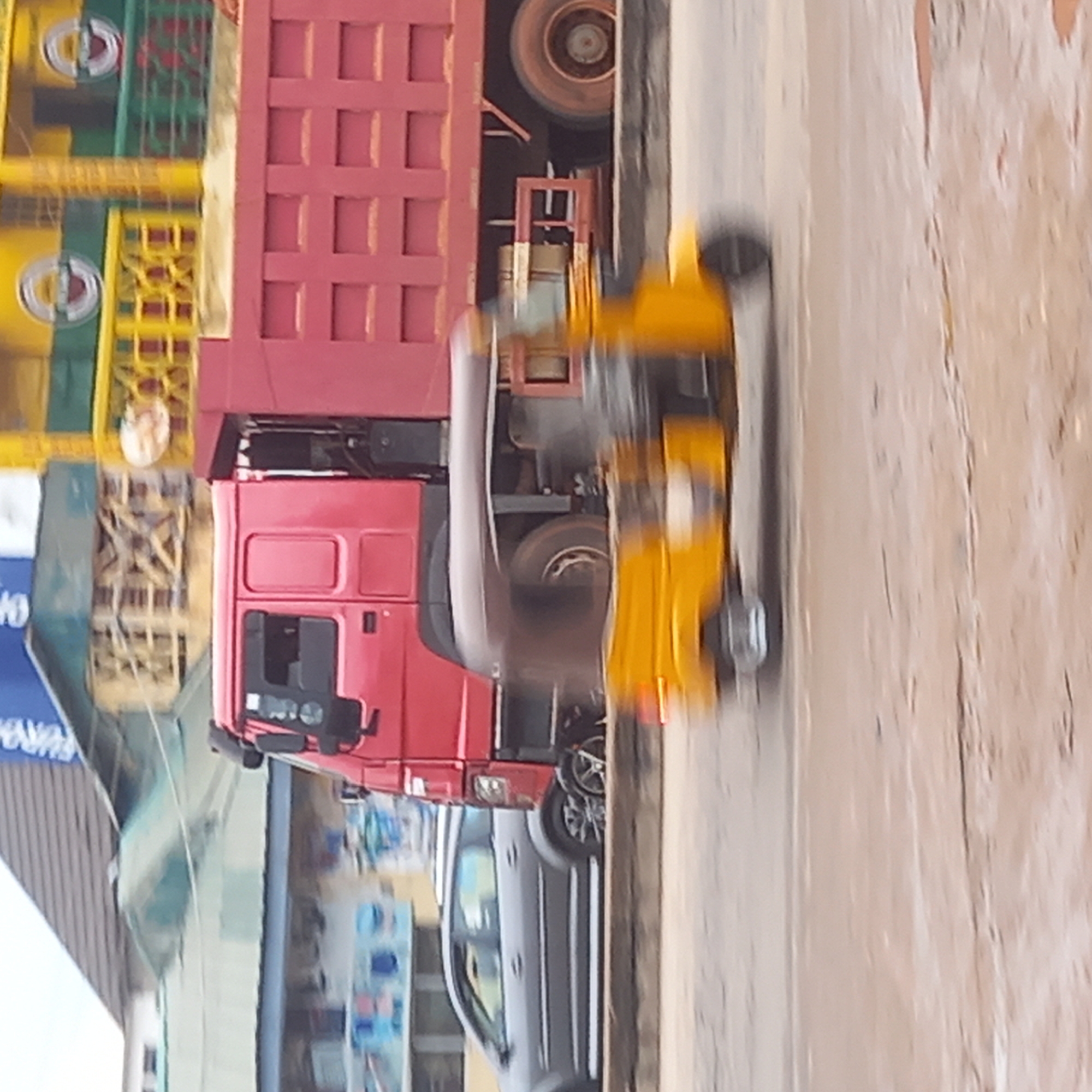 The image depicts a street scene in Dadi Riba, Nigeria. A red truck with a burgundy cargo container is prominent in the center of the frame. To the left of the truck, there are structures, including a building with a sign partially visible. The street surface is visible and appears to be textured. A gray car is seen parked beside the truck. A blurry yellow object is moving along the street next to the truck. The overall scene is somewhat blurred, indicating motion or a low-quality image.