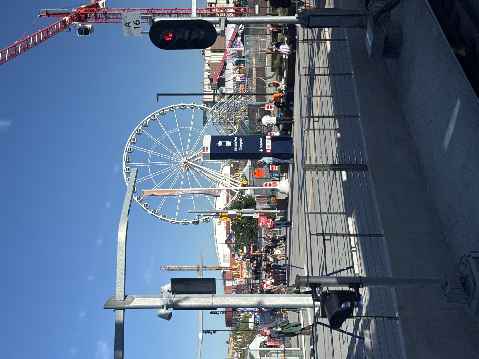 The image depicts a sunny day at the Calgary Stampede in Calgary, Canada. In the foreground, a traffic light with a red arrow is visible, along with a directional sign pointing towards "Victoria Park Stampede" and "Red Line." A Ferris wheel dominates the background, alongside a towering construction crane and various amusement park rides and attractions. People are seen walking and sitting, enjoying the event. The scene is set against a clear blue sky.