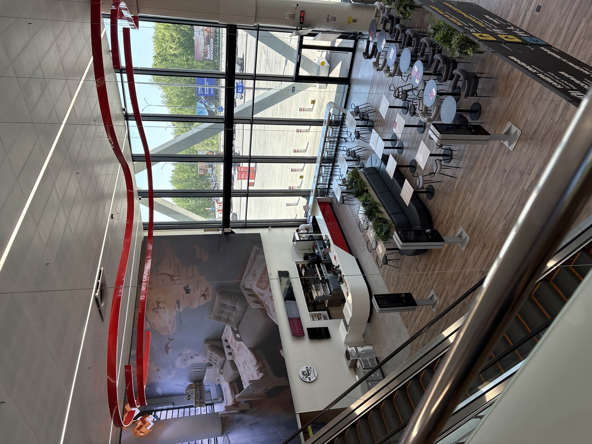 The image depicts an interior space, possibly an airport or large public building, in Новое Мартьяново, Russia, as evidenced by the Cyrillic billboard visible through the window. The space is observed from an elevated position, likely a mezzanine or upper floor, with an escalator in the foreground.
A prominent feature is a large mural on the wall depicting images reminiscent of Russian heritage or history. In front of the mural, there's a modern cafe or bar area with a white counter and seating arrangements. Tables and chairs are arranged in a cafe-like layout, with a mix of round and square tables. Some tables are next to a black cushioned bench.
Large windows offer a view to the outside, revealing trees, buildings, and street signs. A red, decorative ribbon-like structure is suspended from the ceiling, adding a design element to the interior.
The lighting is a mix of natural light from the windows and artificial lighting from the ceiling fixtures. The floor is wooden. The space appears clean and modern, designed to accommodate travelers or visitors.