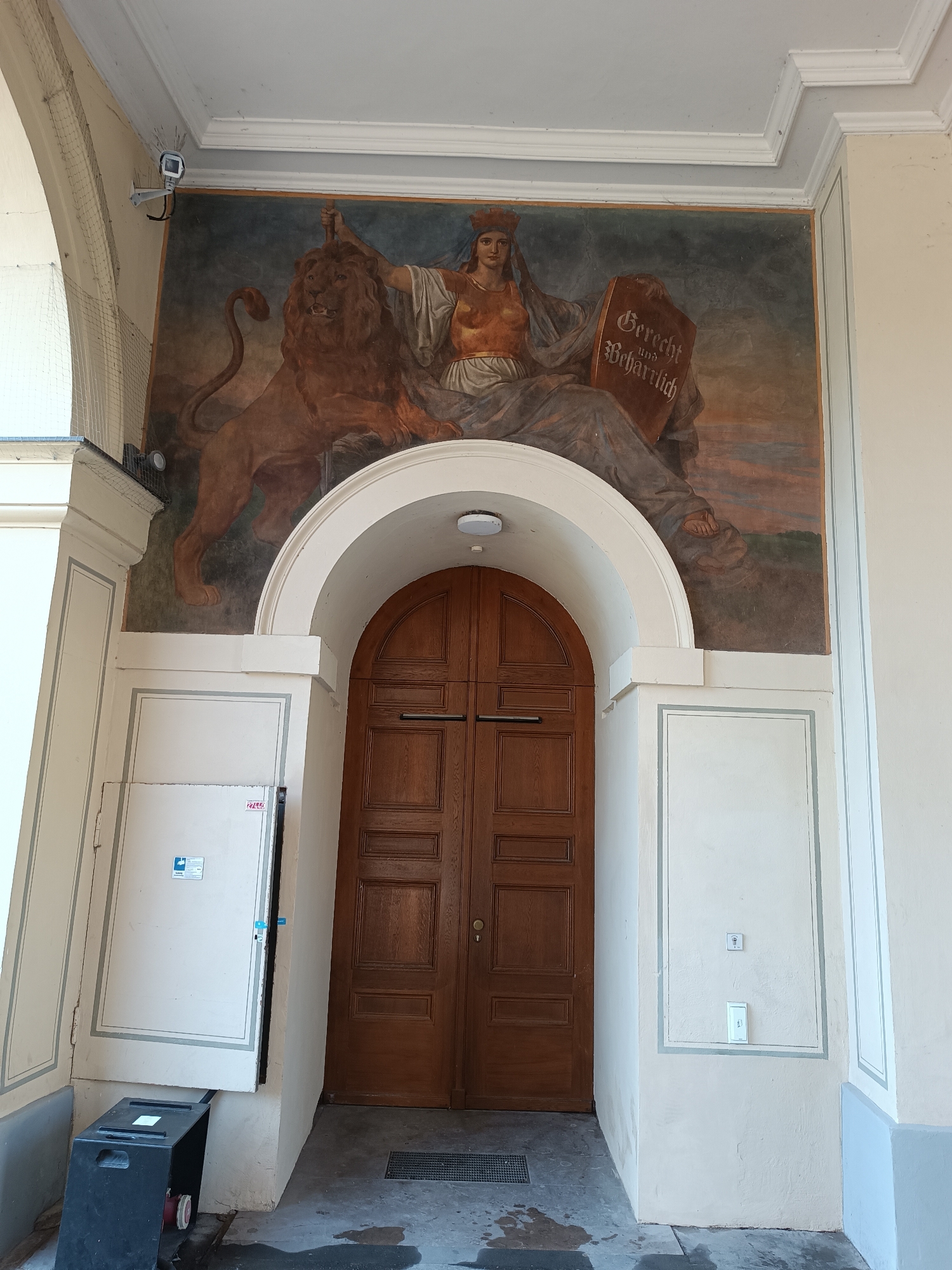 A fresco adorns the exterior wall above a large, arched wooden double door. The fresco depicts a seated female figure wearing a crown and holding a tablet with German text. A lion stands to her left. The scene is set in Munich, Germany, at the entrance of a building. To the left of the doorway, a white electrical panel is visible, and a black bin sits on the ground in front of it. The ground appears wet in some areas.