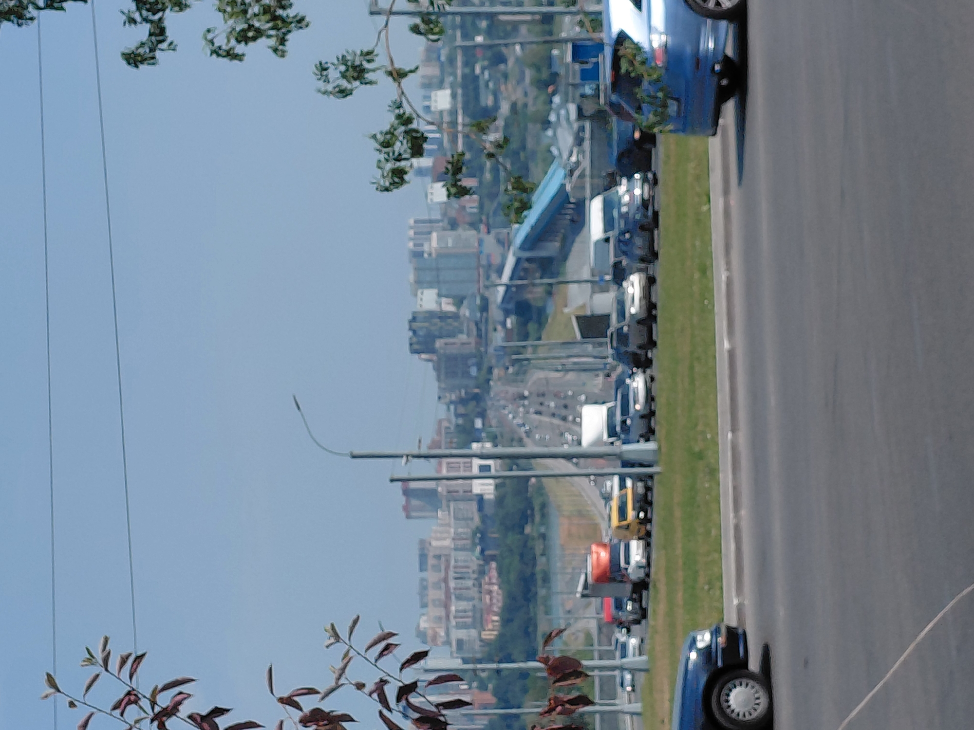 The image shows a panoramic view of a city skyline under a clear blue sky, likely taken from a highway or elevated road. In the foreground, on the right side, a blue SUV is parked or driving. The vehicle appears to be decorated with green foliage around its rear. The SUV is positioned on a paved road, with a grassy embankment behind it.
Further back, a traffic scene unfolds on a multi-lane road, possibly a bridge or elevated highway. Various vehicles, including cars and trucks, are visible. Some vehicles appear to be stopped or moving slowly, indicating traffic congestion. A yellow van is prominent among the vehicles.
In the middle ground, beyond the road, is a body of water, possibly a river, with more urban development on the opposite bank. This is followed by a dense cluster of buildings, suggesting a city center. The architecture of the buildings varies, with some appearing modern and others older. Green trees and vegetation are scattered throughout the cityscape, especially on the hillsides behind the main urban area.
In the background, the skyline is dominated by numerous buildings of different heights and styles. Power lines are visible in the sky. The overall impression is a bustling urban environment with significant infrastructure.
While the image doesn't contain explicit markers to pinpoint Novosibirsk, Russia, the architectural style of the buildings and the general landscape are consistent with many Russian cities, and this type of urban and infrastructure development is common in major Russian cities like Novosibirsk. The presence of a river, hills, and a dense urban center is also characteristic of Novosibirsk.