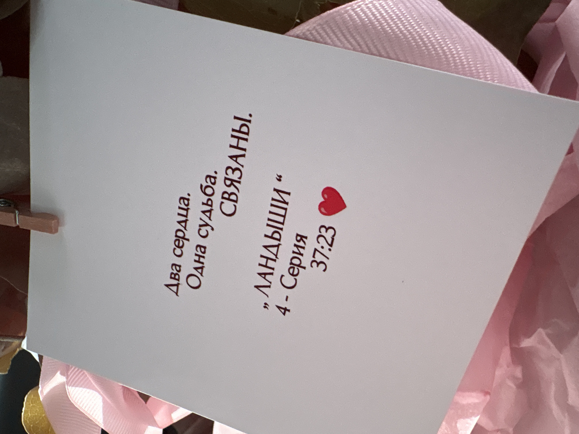 The image displays a white card with Russian text and a red heart icon. The text reads: "Два сердца. Одна судьба. СВЯЗАНЫ. , ЛАНДЫШИ " 4 - Серия 37:23". This translates to: "Two hearts. One destiny. BOUND. , Lily of the Valley " 4 - Series 37:23". The card is partially obscured by pink ribbon and tissue paper, suggesting it might be part of a gift or bouquet. The lighting is soft, and the background is out of focus, with hints of brown and yellow objects, possibly flowers or other decorative elements. The image does not contain any elements that would allow for identification of a specific location in Pavlodar, Kazakhstan.
