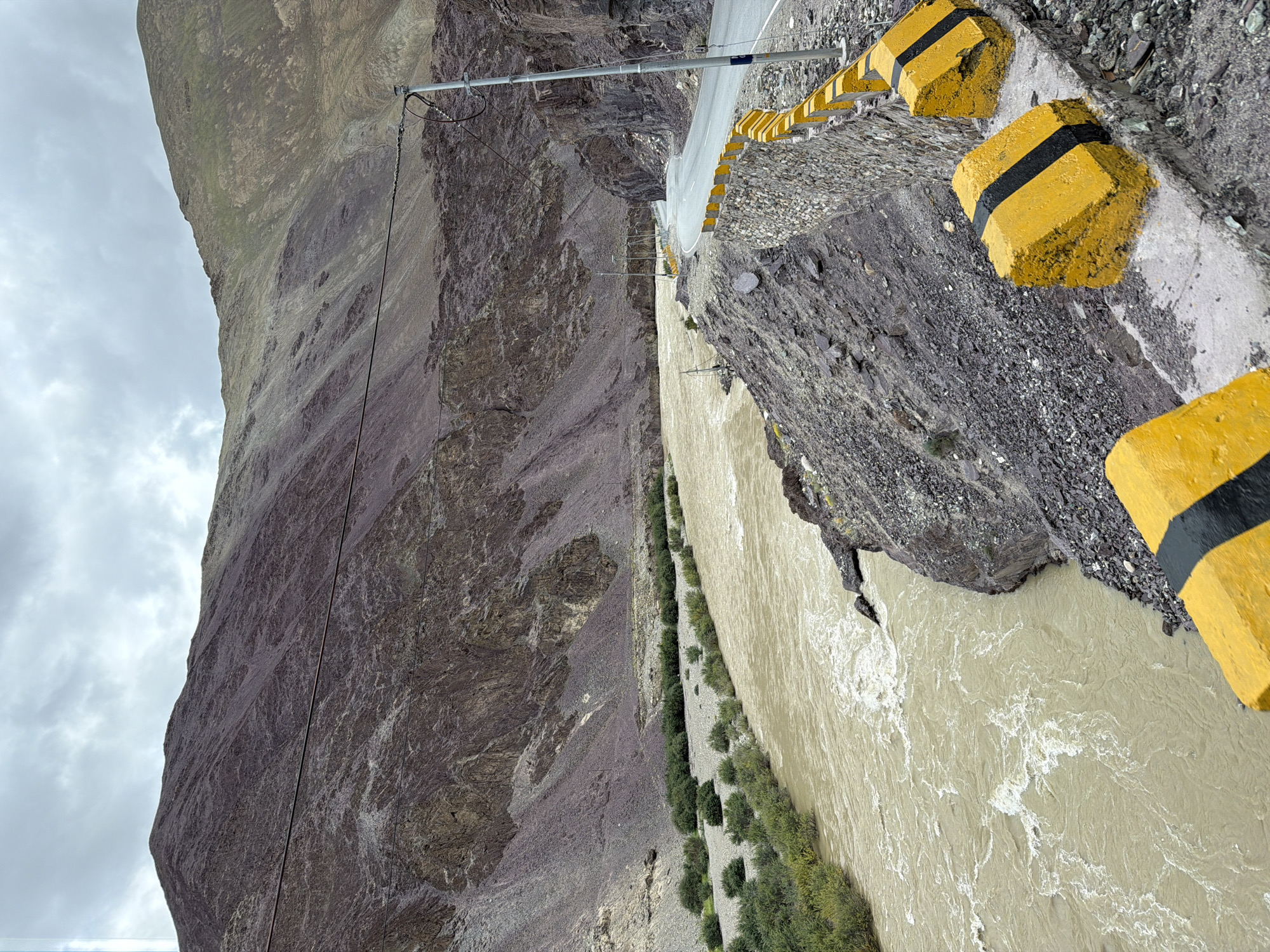 The image depicts a scenic landscape with a powerful, muddy river flowing through a valley. On the right side of the river, a paved road winds its way along the mountainside. The road is bordered by a low, stone wall topped with a yellow and black striped barrier, designed to enhance visibility and safety. Several yellow concrete blocks with black stripes are also positioned along the road's edge. The mountains on either side of the valley are rugged and rocky, with sparse vegetation. The sky is overcast, with a grey, cloudy formation. The scene appears to be captured in a remote, mountainous region, likely in or around Kiari, India, given the context. There are no people or animals visible in the image. The overall impression is one of a dramatic, natural environment shaped by the elements and traversed by human infrastructure.