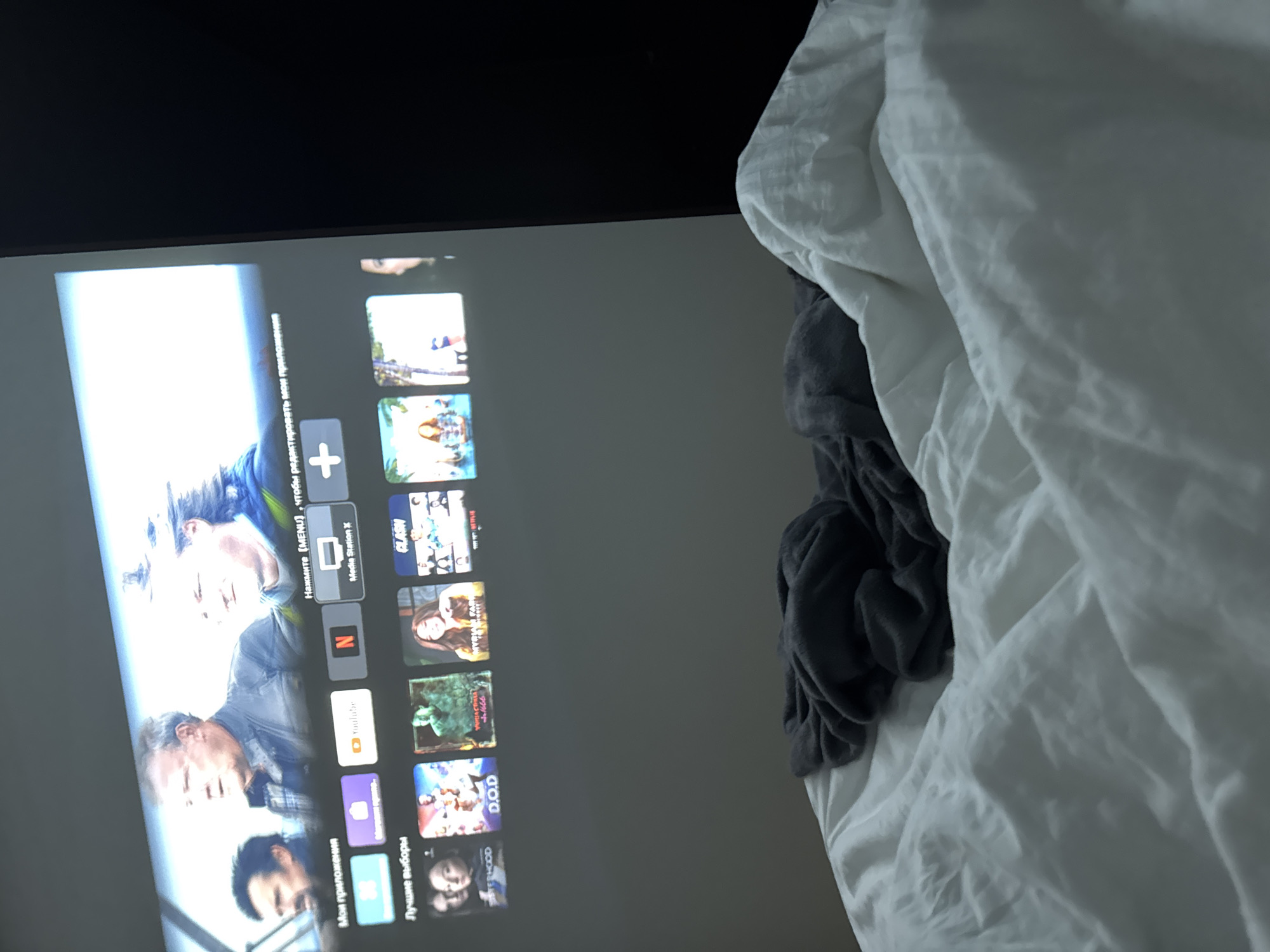 The image shows a television screen displaying a menu of applications. The screen is situated beside a rumpled white bedsheet and a gray blanket. The applications visible on the screen include YouTube, Netflix, and Media Station X. The title "Мои приложения" (My Applications) and "Лучшие выборы" (Best Choices) are displayed, indicating this is a smart TV interface.