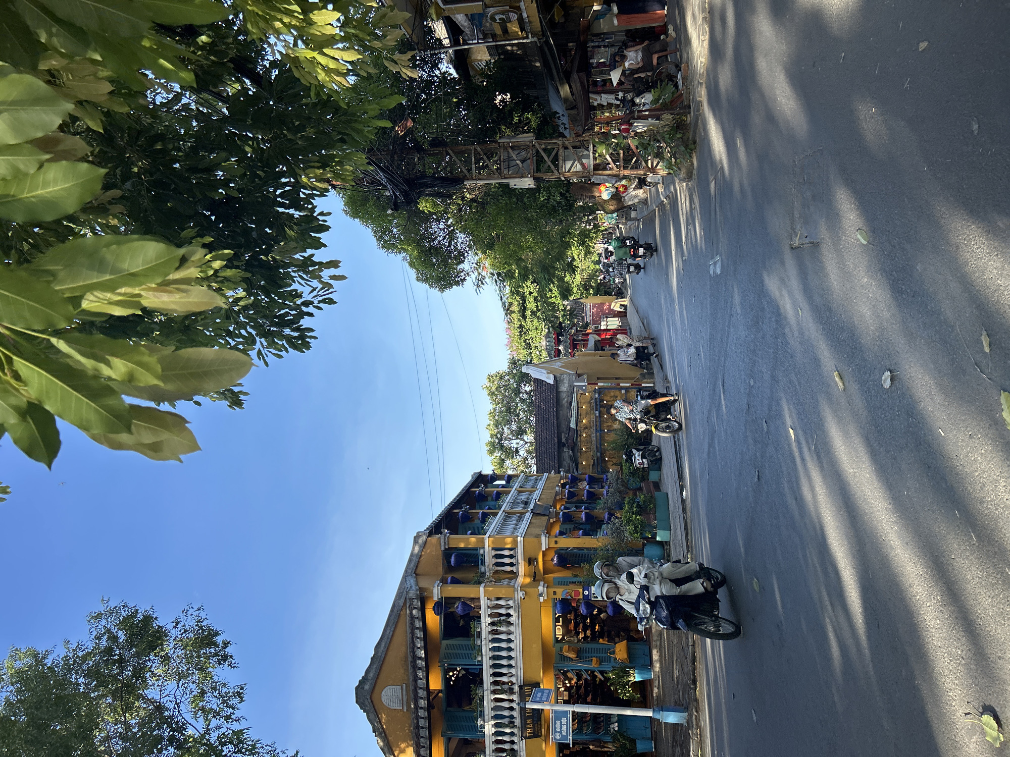 The image depicts a sunlit street scene in Hội An Ward, Vietnam, characterized by traditional architecture and active street life.
In the foreground, an asphalt road displays distinct, elongated tree shadows from the left. A dark scooter carrying two helmeted individuals in light-colored clothing is in motion in the left lane, traveling away from the viewer. To its right, a lone rider on a yellow motorbike also travels in the same direction. Scattered dry leaves are visible on the road surface.
On the left side of the street, a prominent two-story building painted bright yellow features a white balustraded balcony on the second floor, adorned with multiple blue-purple lanterns. The windows are fitted with blue louvred shutters. The ground floor appears to be a shop, with footwear and handbags visible through the open front. A blue street sign post near the building reads "THAN THƯƠNG !!!" and "ĐƯỜNG HOÀNG DIỆU". Potted plants in blue planters line the sidewalk.
The right side of the street features a dense network of electrical wires attached to a tall, weathered metal utility pole, partially obscured by lush green trees. Further down the street, several smaller shops and buildings line the road. A group of individuals is seated on low stools by the roadside under an awning; one person wears a traditional conical hat (nón lá). Multiple motorbikes are parked along the curb and several others are in motion further down the street.
The sky is clear and bright blue, visible above the buildings and through the lush green foliage that frames the upper corners of the image. The strong sunlight indicates a daytime setting.