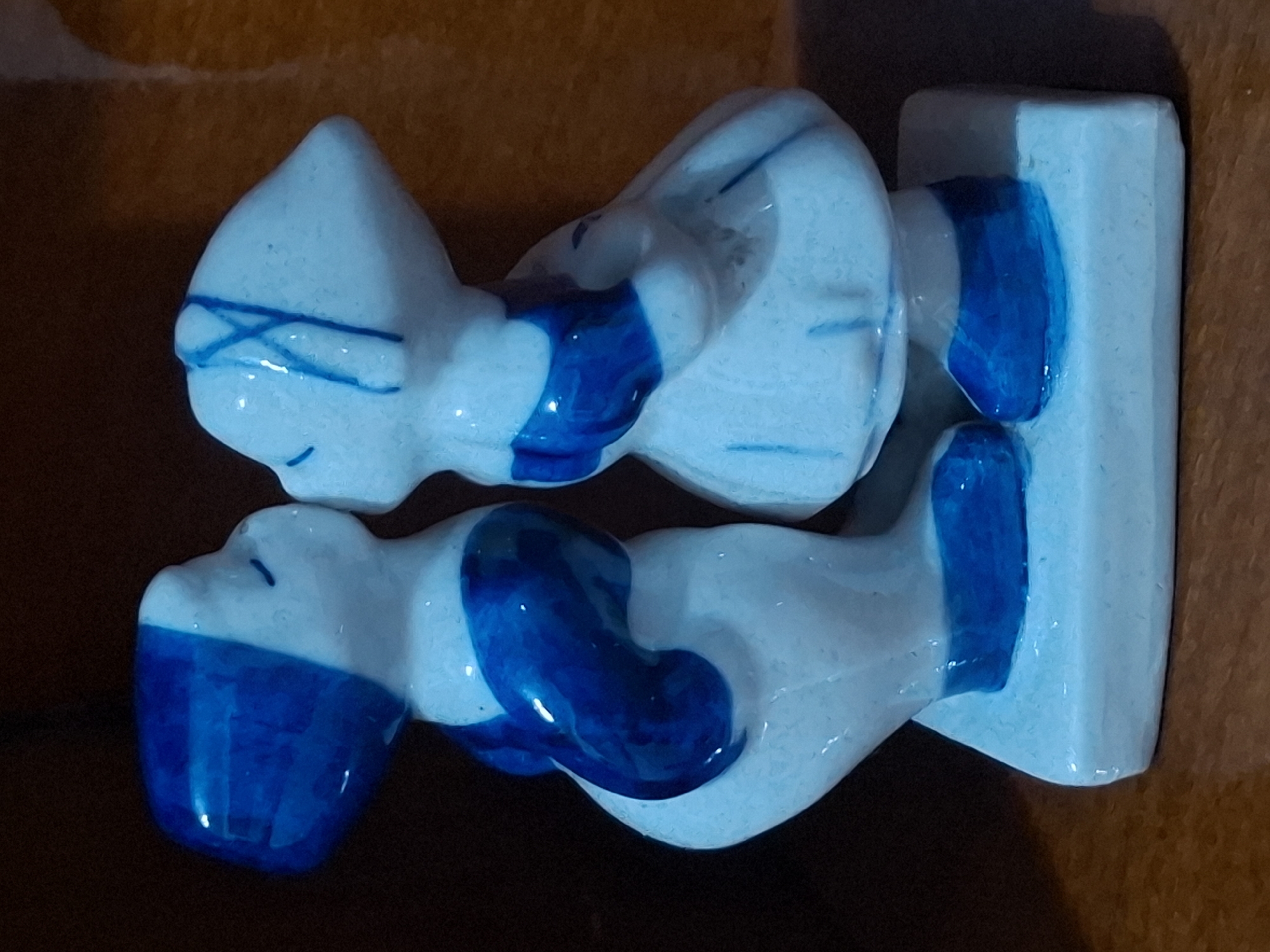 A close-up, eye-level, medium shot captures a ceramic figurine of two children, a boy and a girl, depicted in the act of kissing. The figurine, predominantly white with blue accents, stands on a light blue rectangular base. The boy, on the left, wears a blue cap and a blue garment. The girl, on the right, has blue lines suggesting hair ties and wears a blue-accented dress. Both figures have their eyes closed, leaning towards each other to share a kiss. The figurine is set against a dark, out-of-focus background with hints of warm brown tones, suggesting it is placed on a wooden surface. The lighting is diffused, highlighting the glossy finish of the ceramic. This image is not associated with the city of Almaty, Kazakhstan.