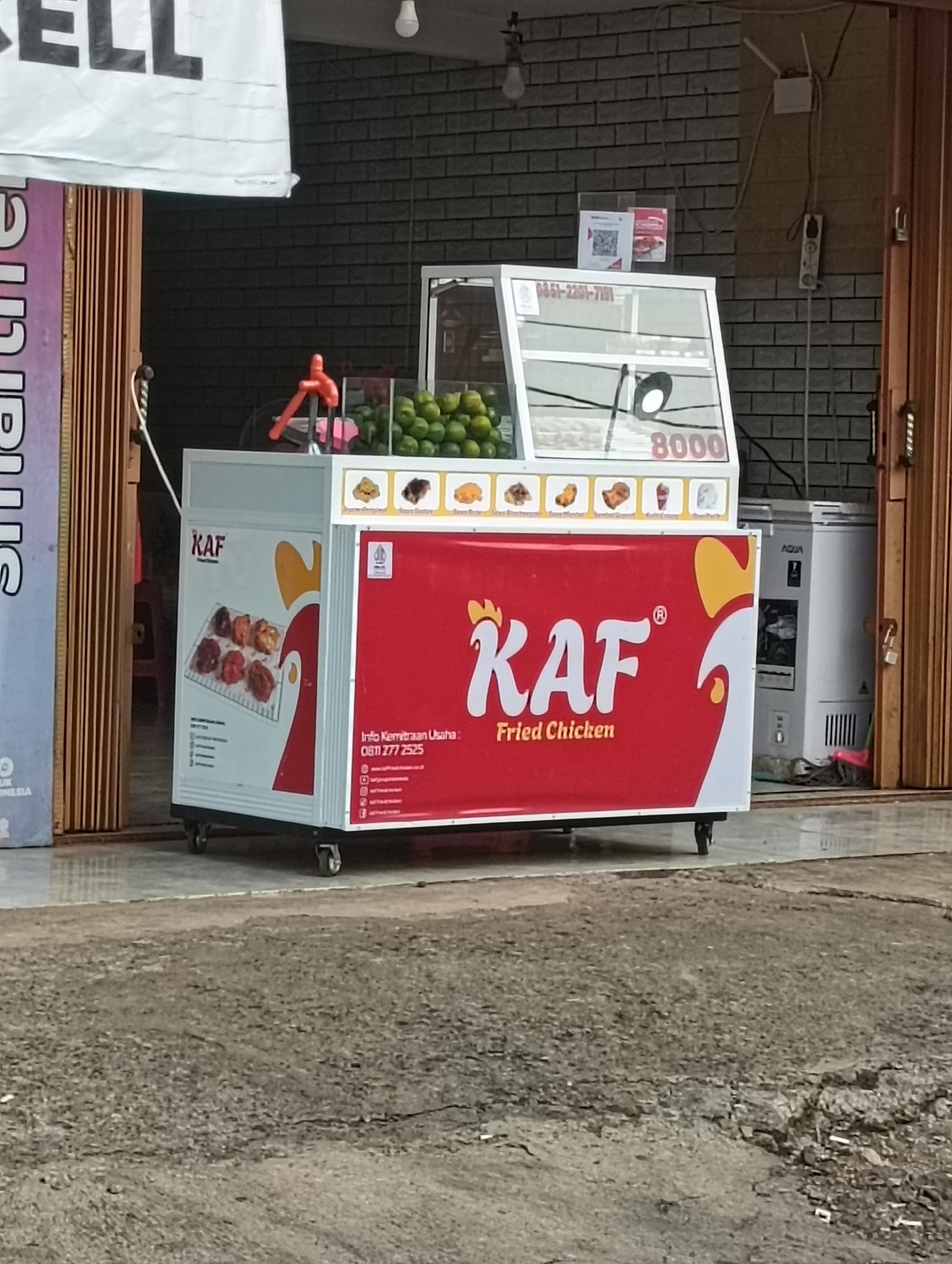 In Tayu, Indonesia, a food cart is displayed outside a store. The cart features a red body with "KAF Fried Chicken" written in large white letters, accented by yellow chicken graphics. On the side, a picture shows pieces of fried chicken. Above the food cart, a glass display case showcases fresh green limes. Next to the cart, a white refrigerator is visible, and a rolling metal door is partially open, revealing a brick wall behind it. The ground in front of the cart is paved with cracked concrete, and a white banner with the word "SELL" in large black letters hangs above the store entrance.