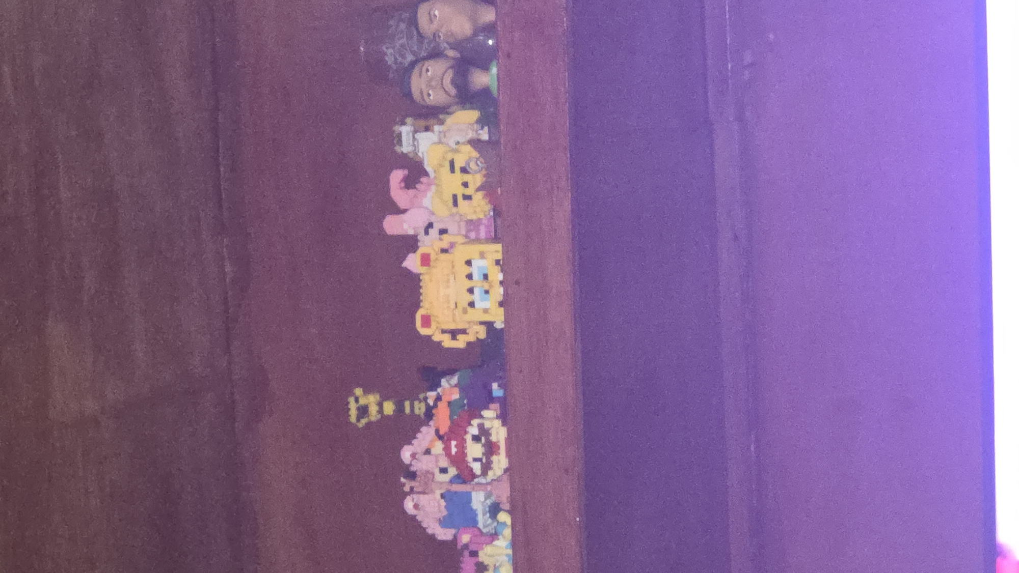 The image shows a shelf with various pixel art figures and a few busts on display. From left to right, there is a yellow figure with a spiky hairdo, followed by a figure that appears to be Super Mario. Next, there is a yellow square figure with blue eyes, resembling SpongeBob SquarePants. To its right, there are some pink figures, and further right, two male busts with dark hair and beards. The shelf is made of wood and is mounted on a wooden wall. The background is a dark, textured surface, possibly wood paneling. The lower portion of the image is blurred and shows a purple surface, likely a table or floor. The lighting is dim, casting shadows on the objects. The scene is indoors, likely in a domestic setting or a display area. There is no visible indication of the location in East Jakarta, Indonesia.