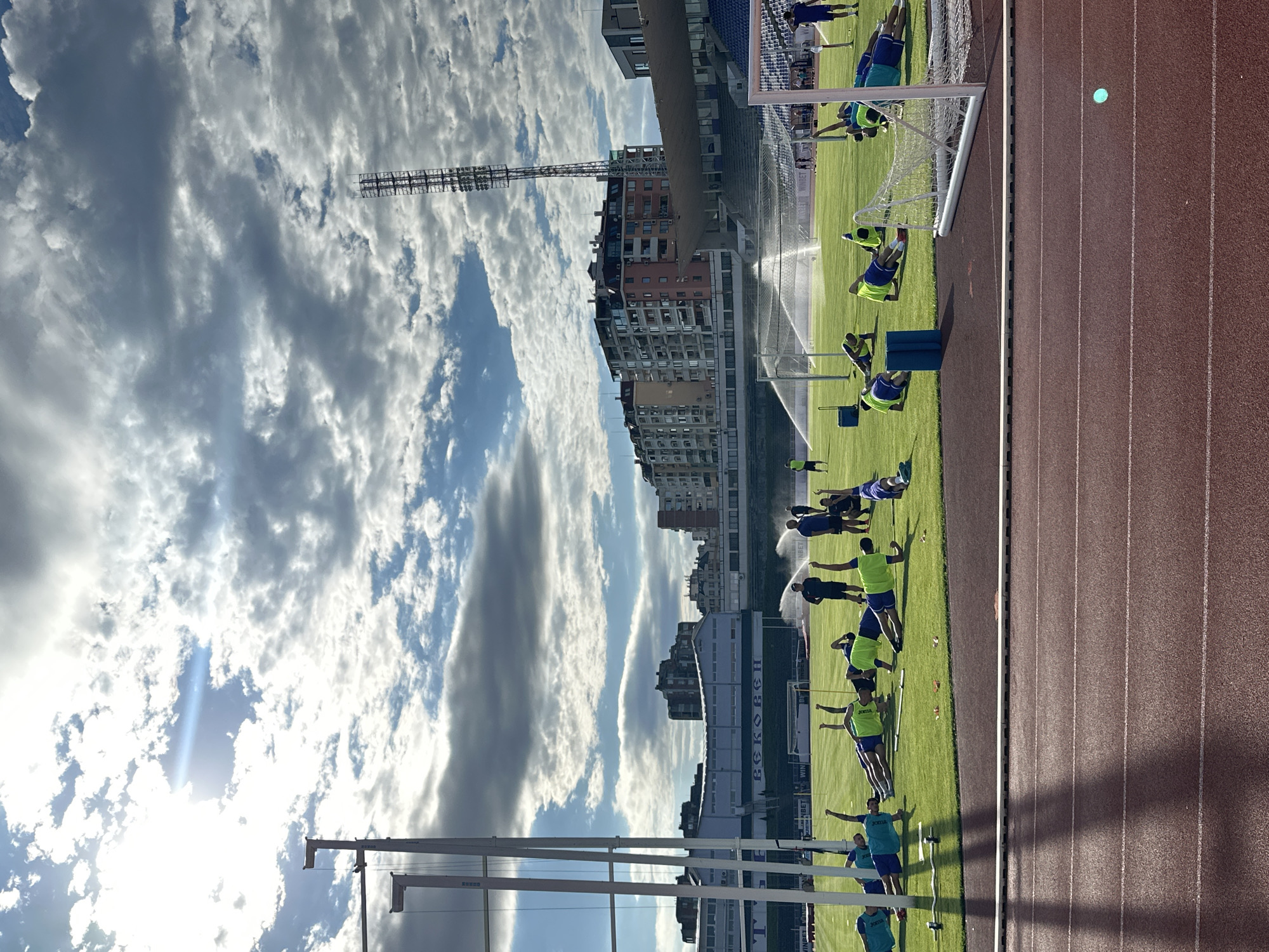 The image shows an outdoor training session on a grassy field at the Ivaylo Stadium in Veliko Tarnovo, Bulgaria. A group of athletes, wearing blue and green sportswear, are performing exercises, primarily planks, on the field. Some of the athletes are being supervised by coaches, two of whom are wearing dark uniforms and standing near a water sprinkler system that is actively watering the field. In the background, there are multi-story buildings, stadium seating, and a tall light tower. The sky is partly cloudy with the sun shining through, creating a dramatic lighting effect. The foreground shows a portion of a red running track.